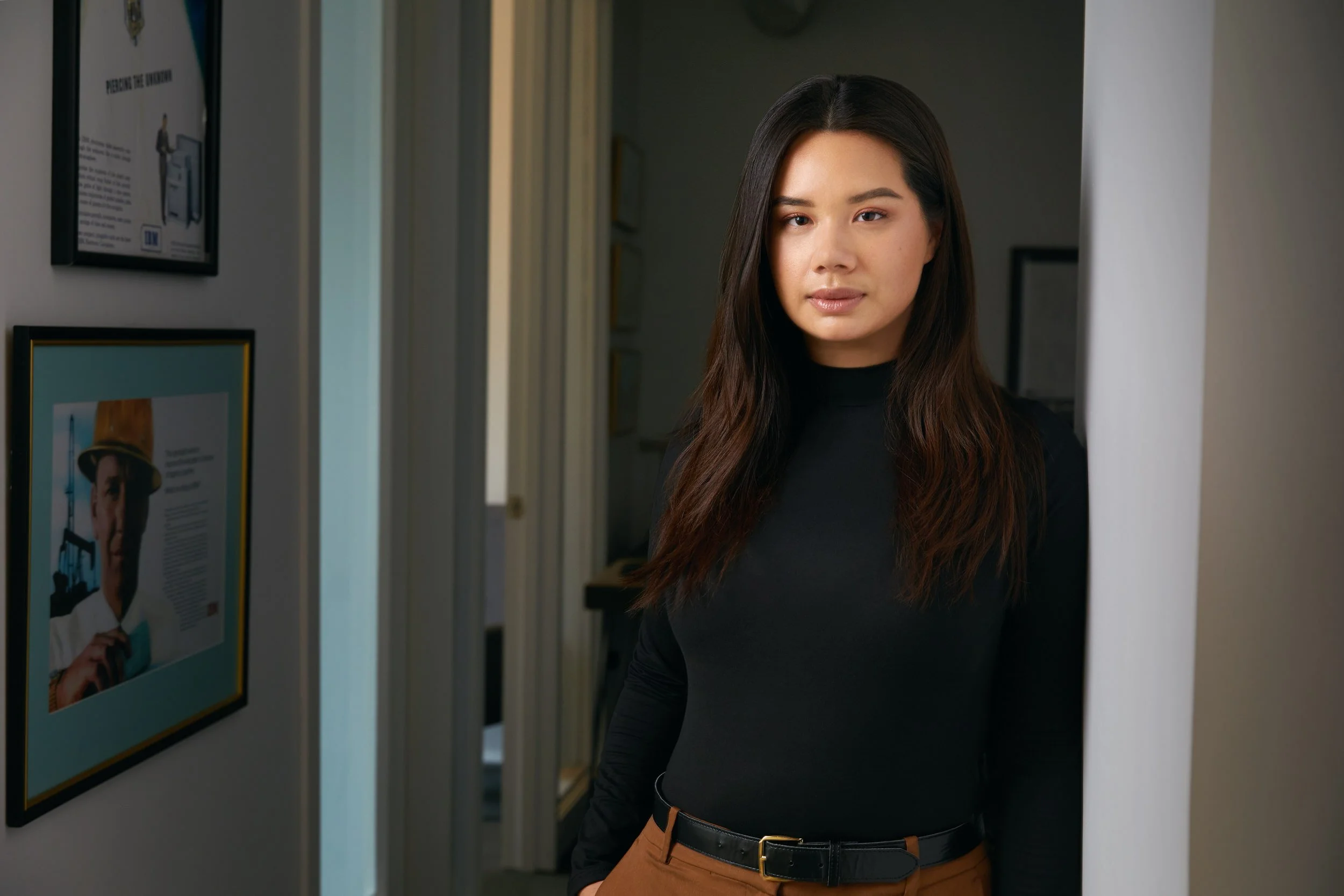 Office portrait standing in a doorway, black sweater, framed prints on the wall