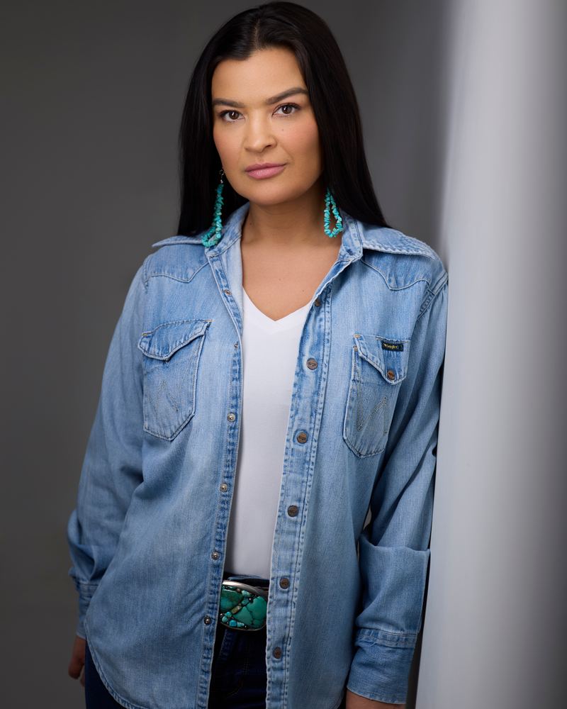 Crystle Lightning in a denim shirt over white, beside a pale wall