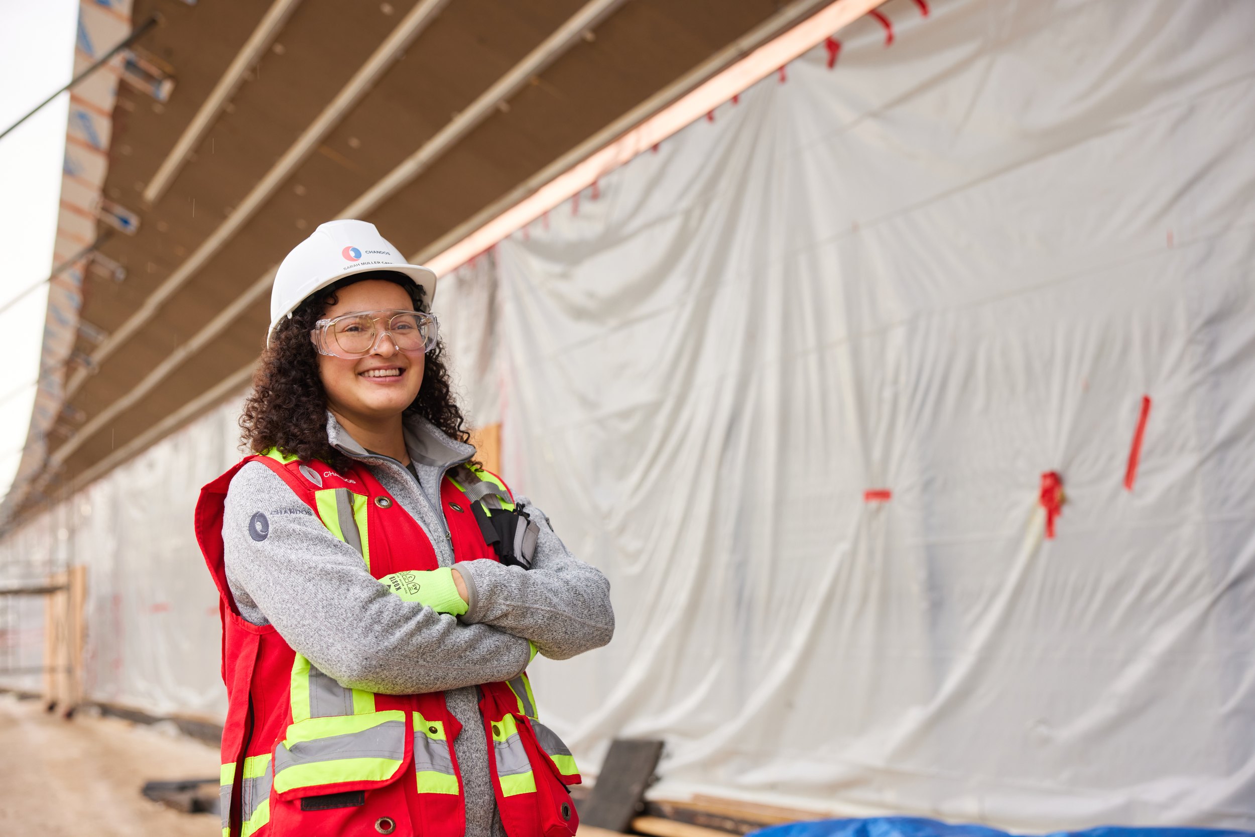 A woman in a hard hat and vest, arms crossed, by a tarped wall