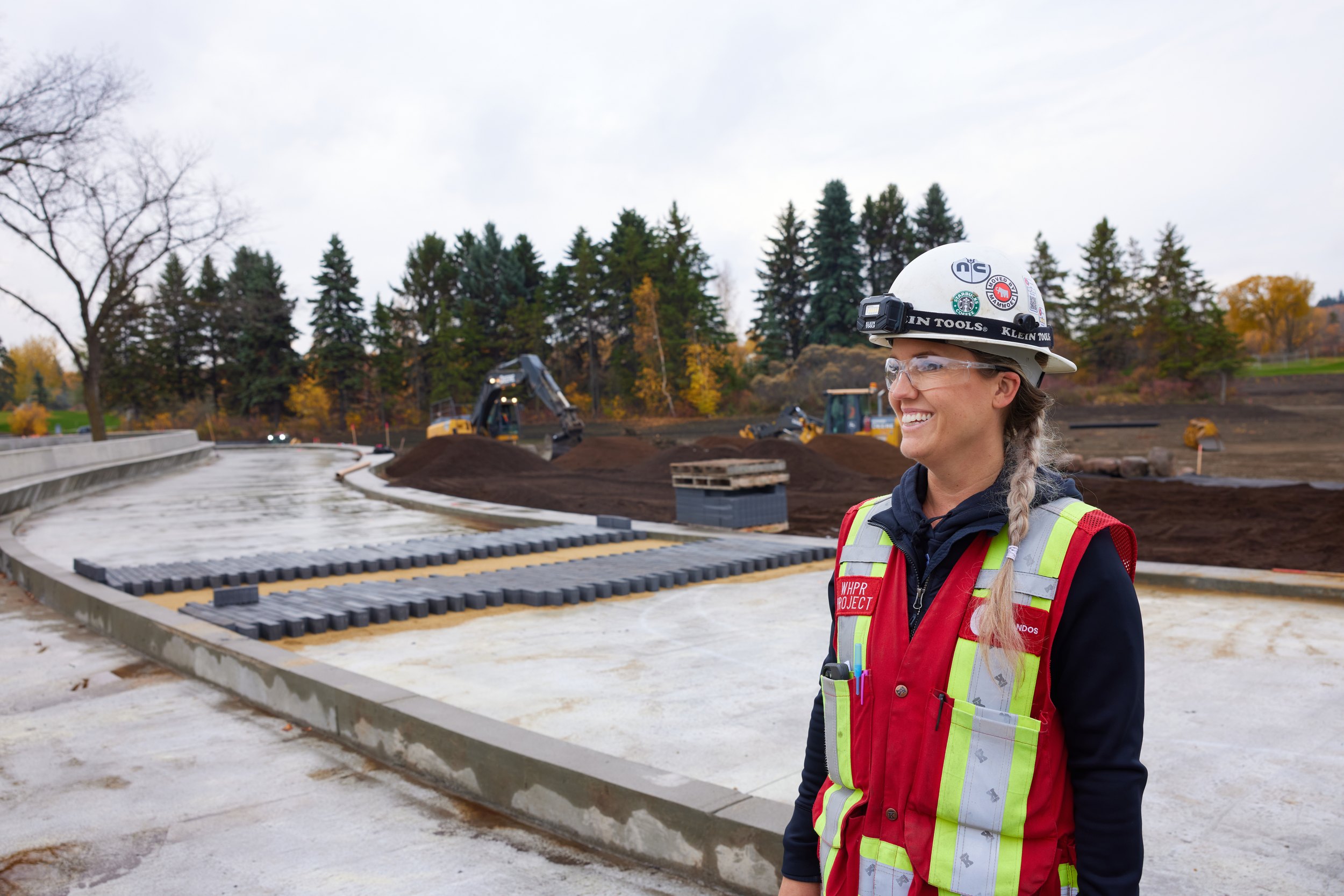 A woman in a hard hat beside a curve of fresh concrete, trees behind