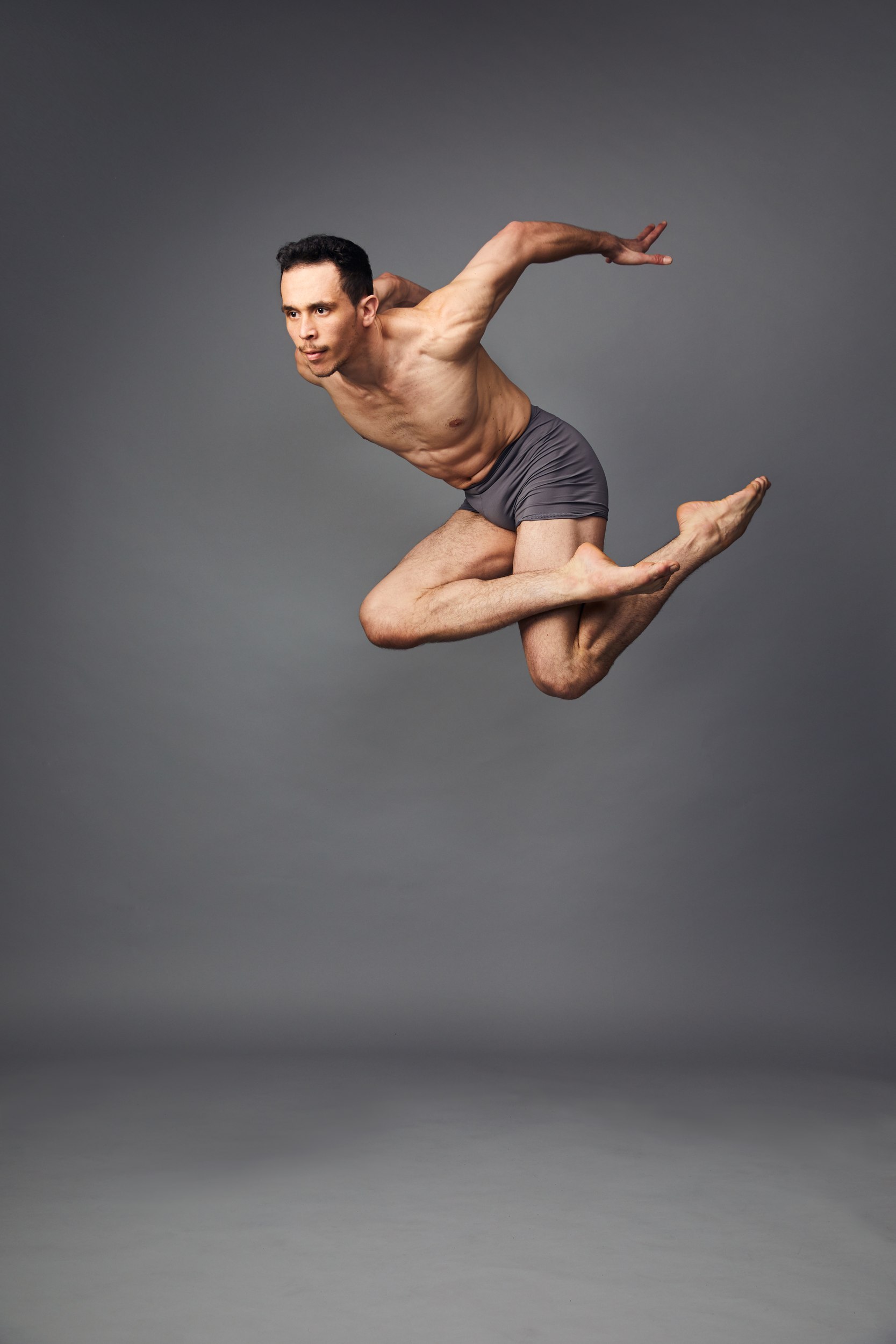 A dancer in grey shorts mid-leap on grey, knees bent under him, one arm swept back