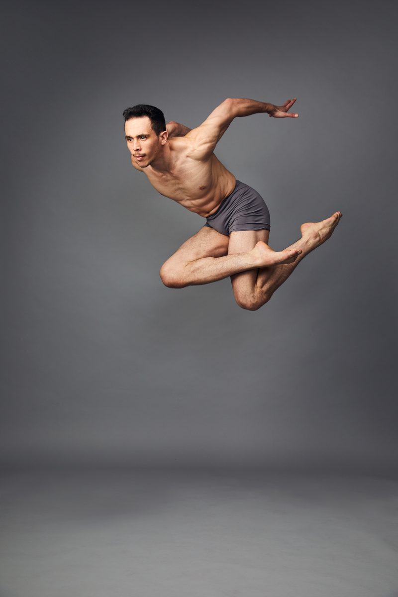 A dancer in grey shorts mid-leap on grey, knees bent under him, one arm swept back