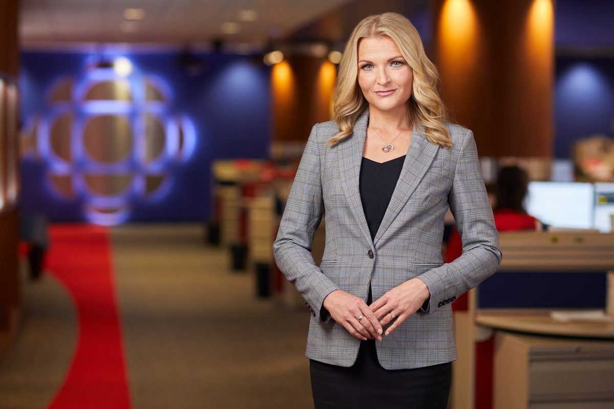 A broadcaster standing in a network newsroom, the desk behind her