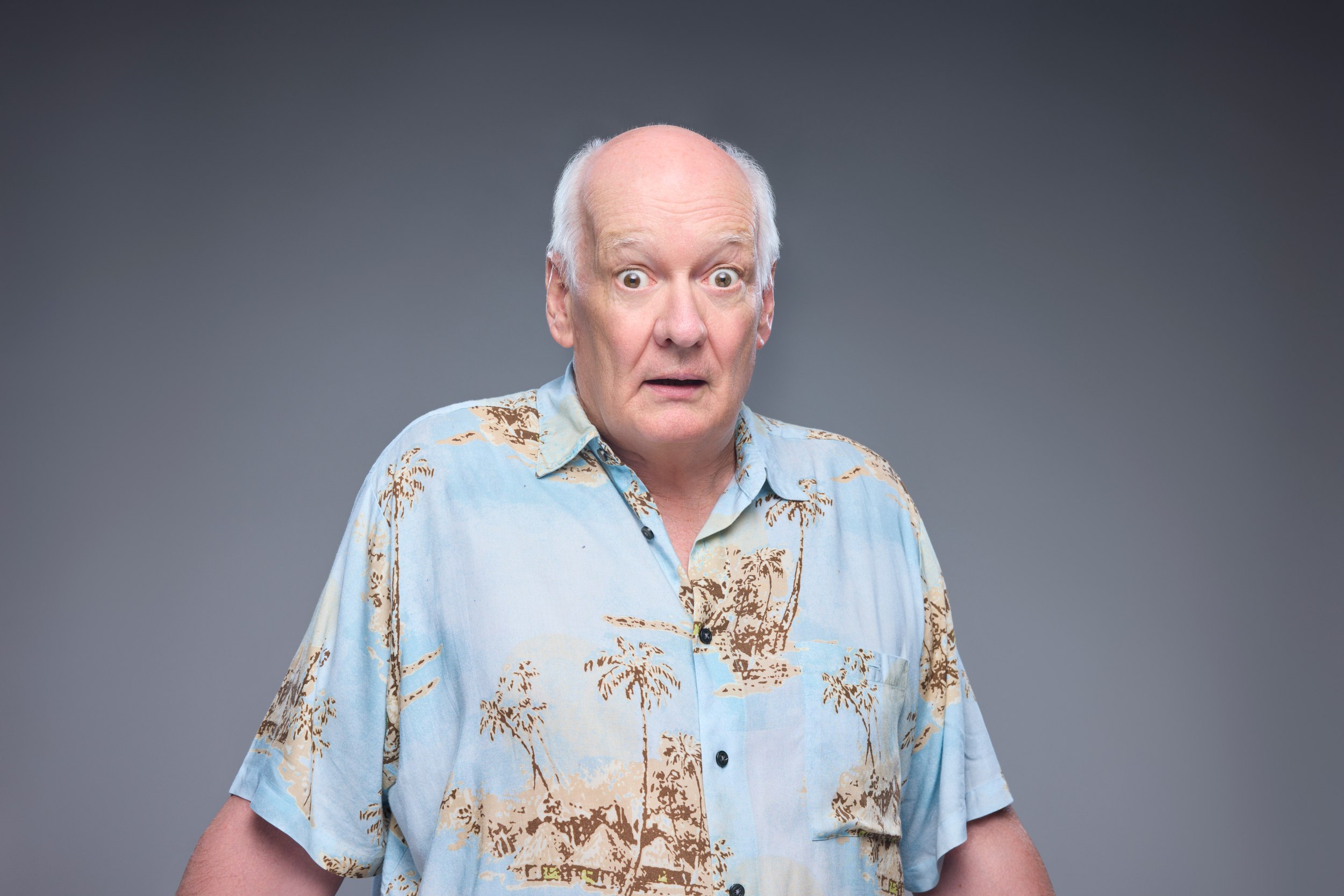 Colin Mochrie in a palm-print shirt, eyes wide, against grey