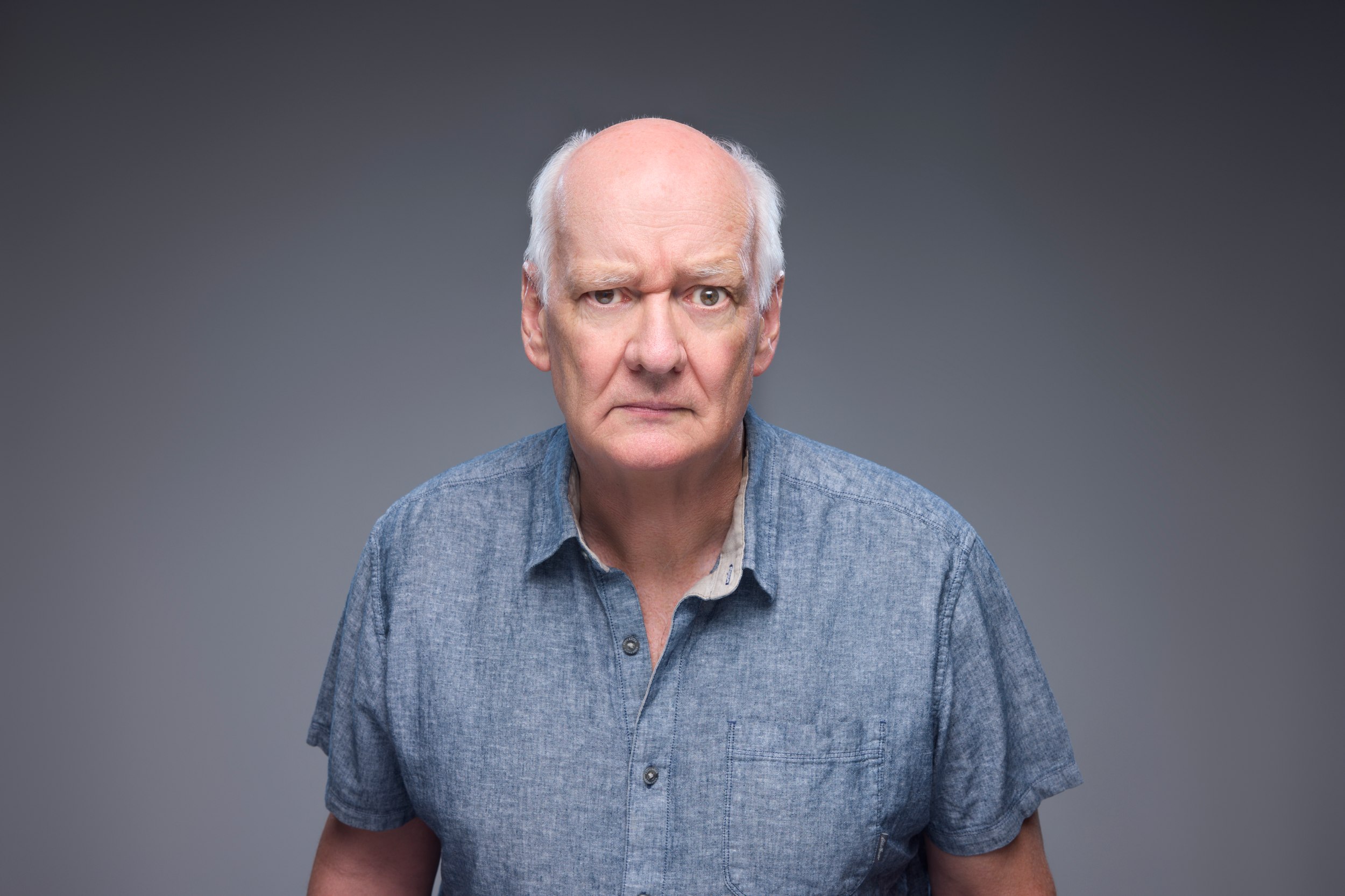 Colin Mochrie in a blue linen shirt, a stern look, against grey