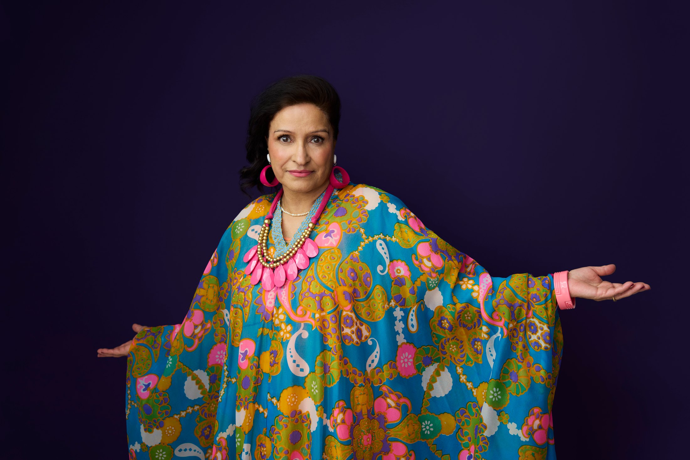 A woman in a bright printed kaftan, arms wide, against deep blue