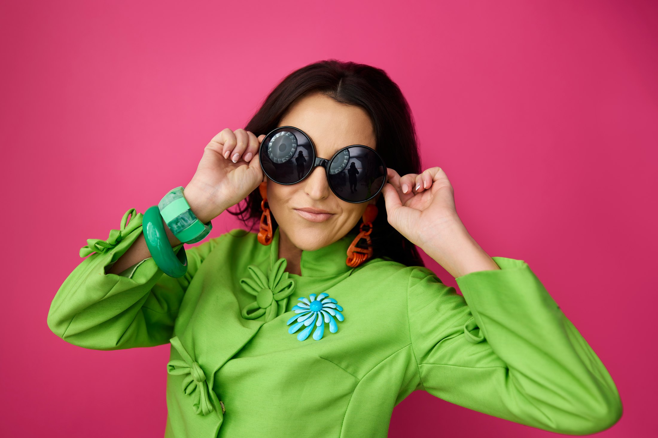 A woman in a green top lowering her sunglasses, against magenta