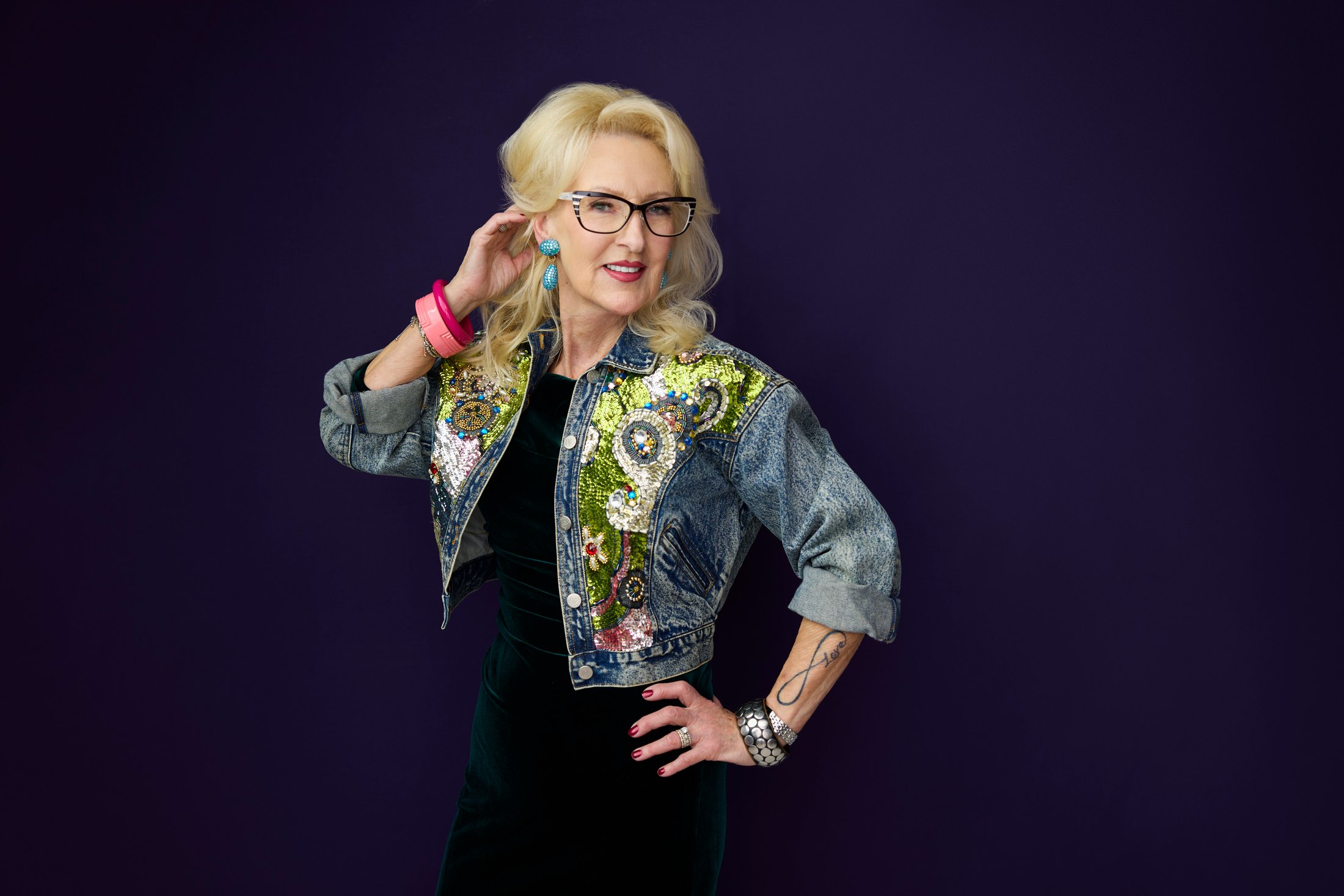 A woman in a patterned jacket, a hand on her hip, against violet