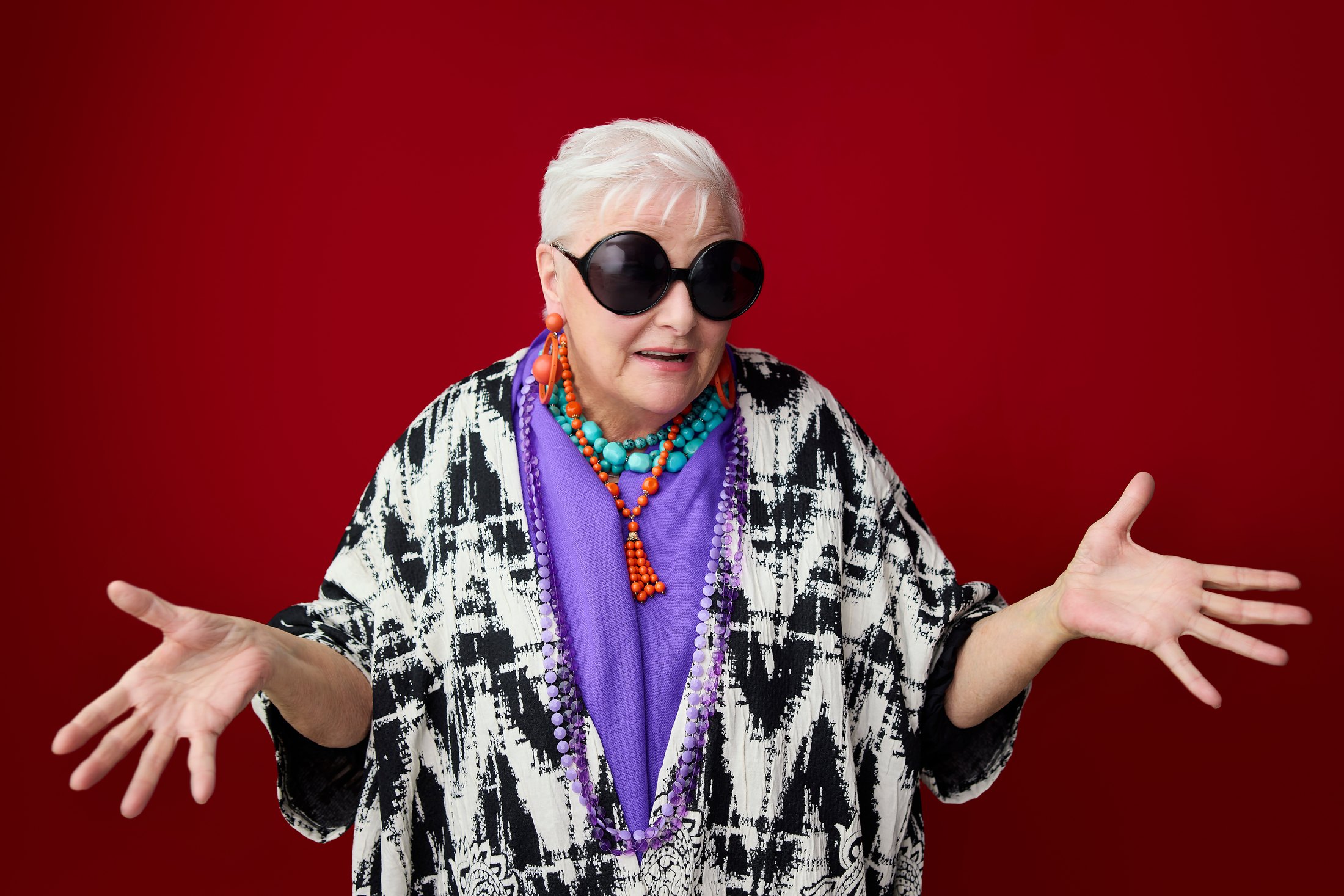 A woman in a monochrome print coat and sunglasses, hands out, against red