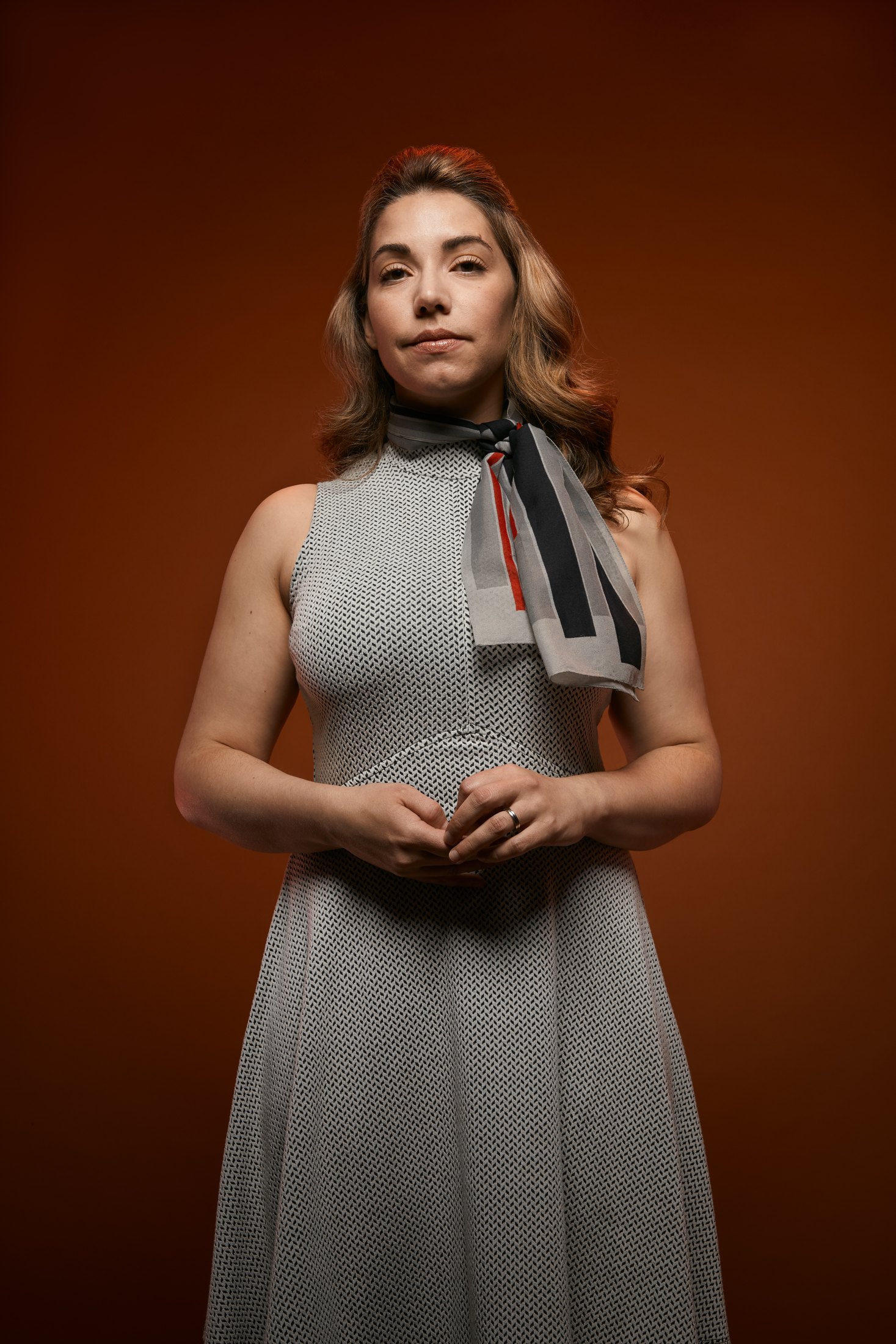 A woman in a grey halter dress and neck scarf, hands clasped, against rust