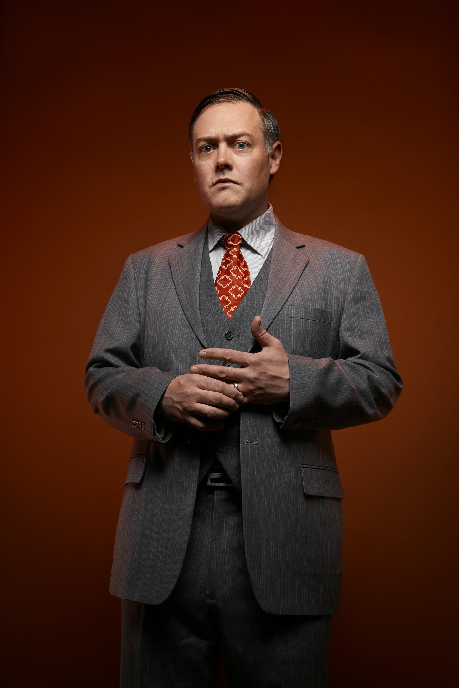 A man in a grey suit and patterned tie, hands clasped, against rust