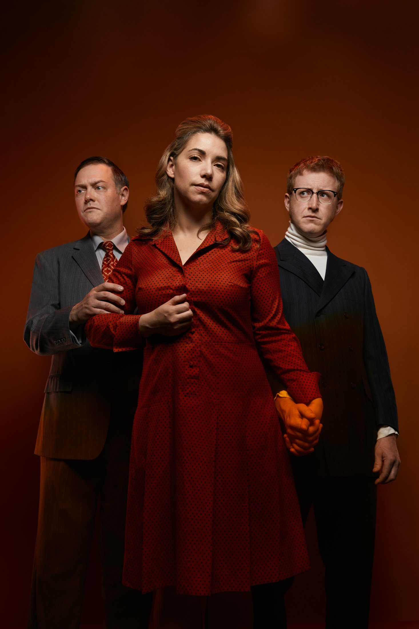 Three performers against rust, a woman in red between two men in suits