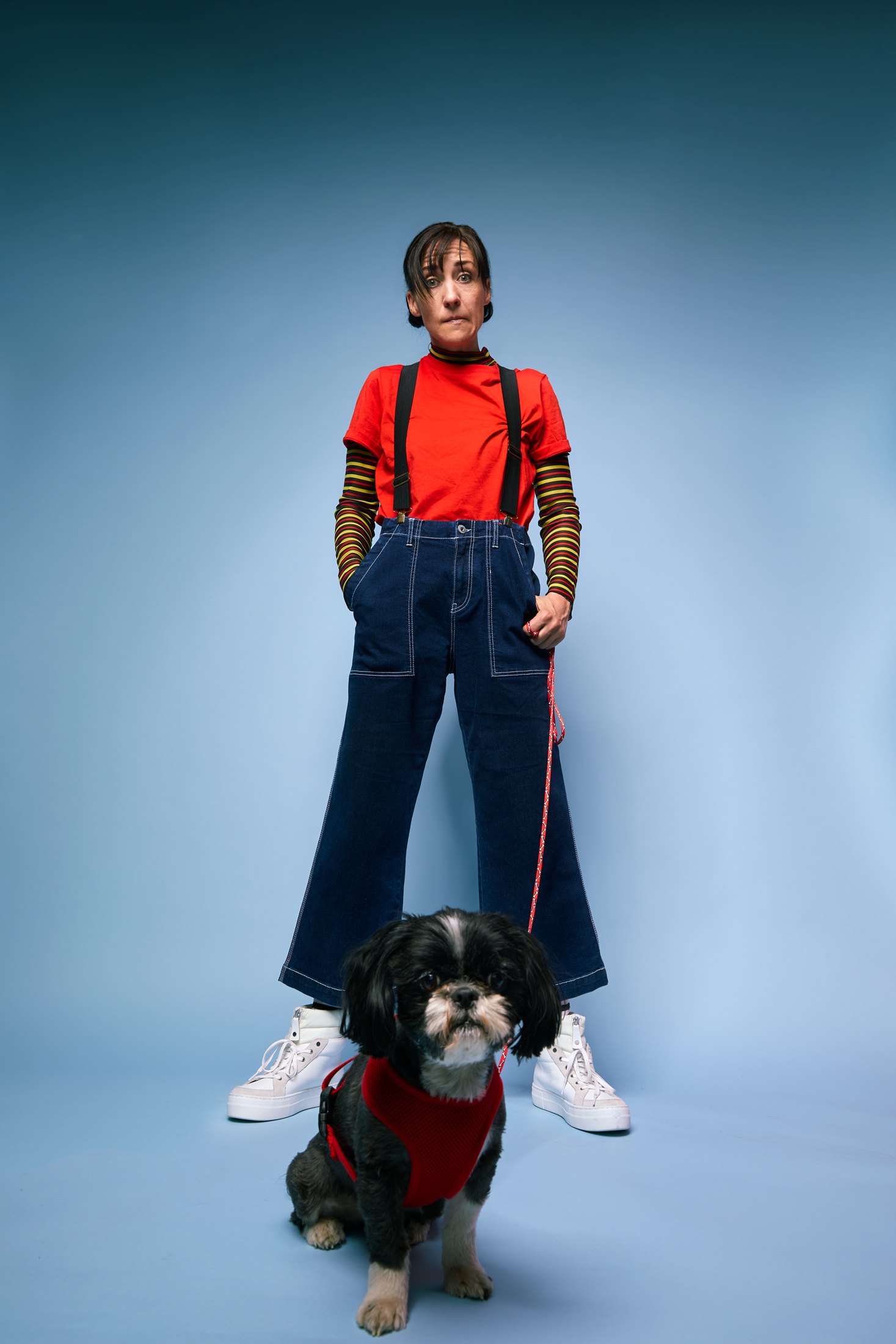 A performer in a red top and denim overalls standing over a small dog