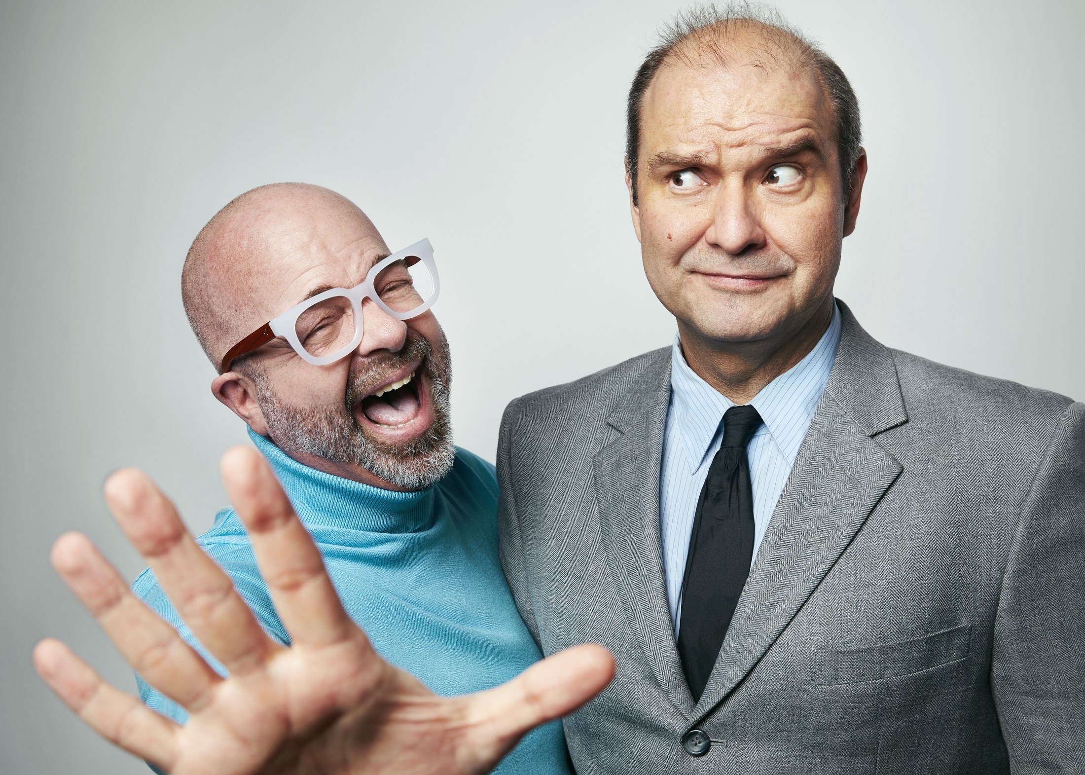 Two performers on pale grey, one in a turquoise turtleneck and pink glasses with a hand out, one in a grey suit