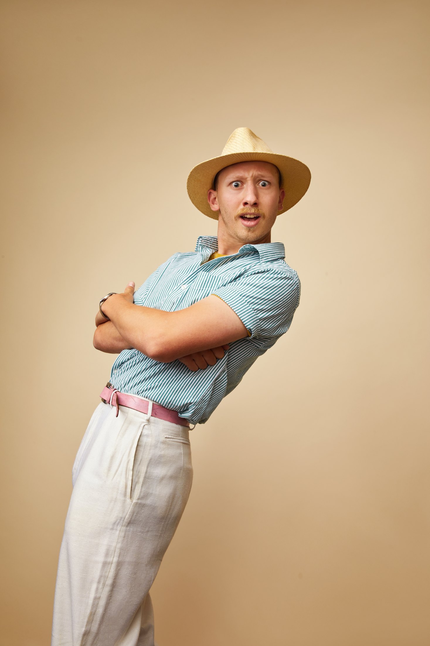 A performer in a straw hat and pale blue shirt, arms crossed, against tan