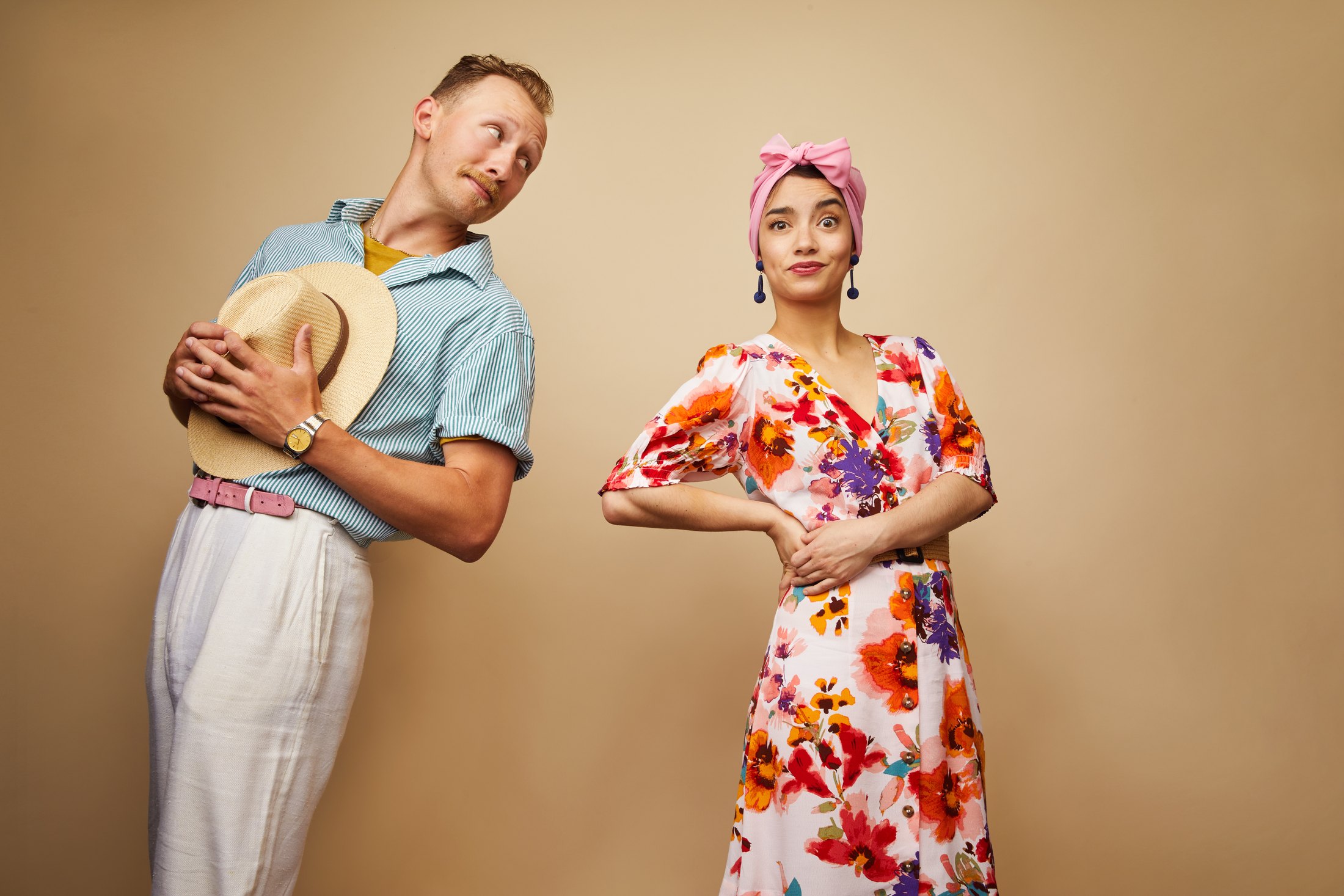 Two performers against tan, one holding his straw hat, one in a floral dress and headwrap