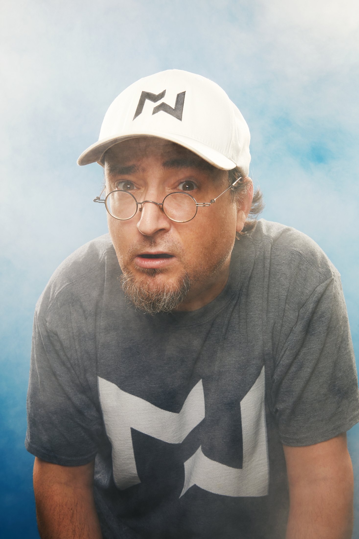 A performer in a white cap, glasses and a printed tee against pale blue