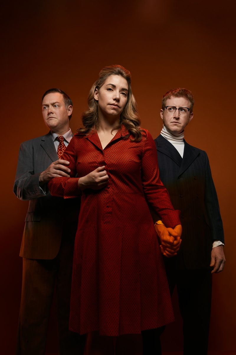 Three performers against rust, a woman in red between two men in suits