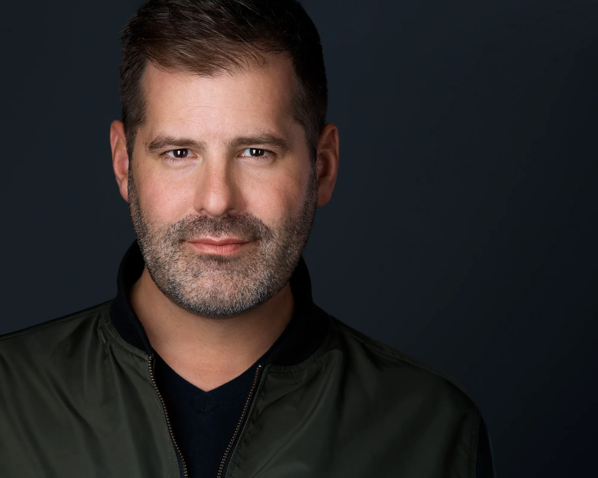 A broadcaster in an olive bomber jacket against near-black, square to the camera