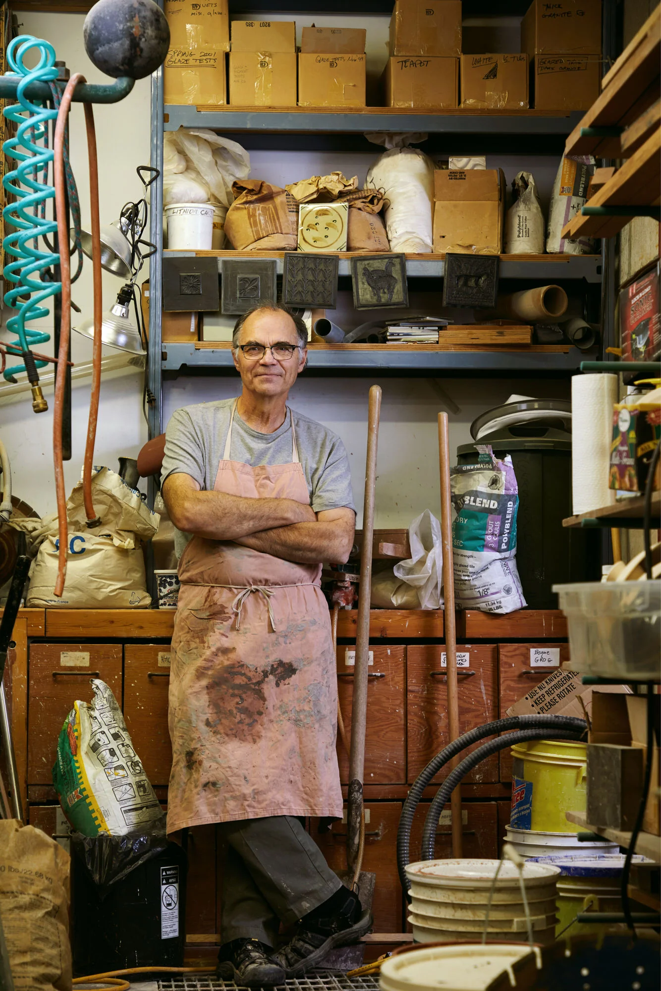 A potter in a clay-stained apron, arms crossed, among the shelves of his workshop