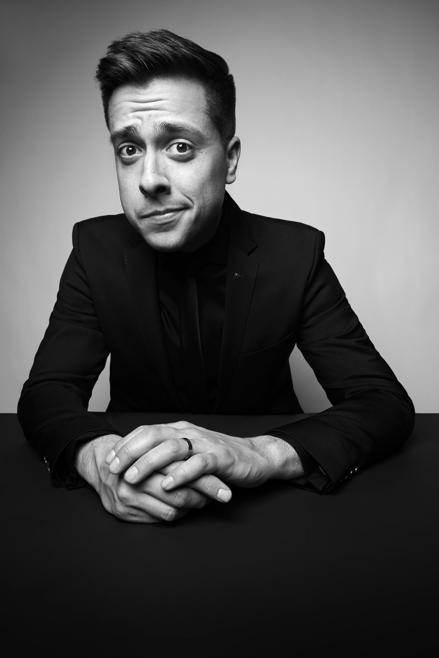 In black and white, a dark jacket, hands folded on the table
