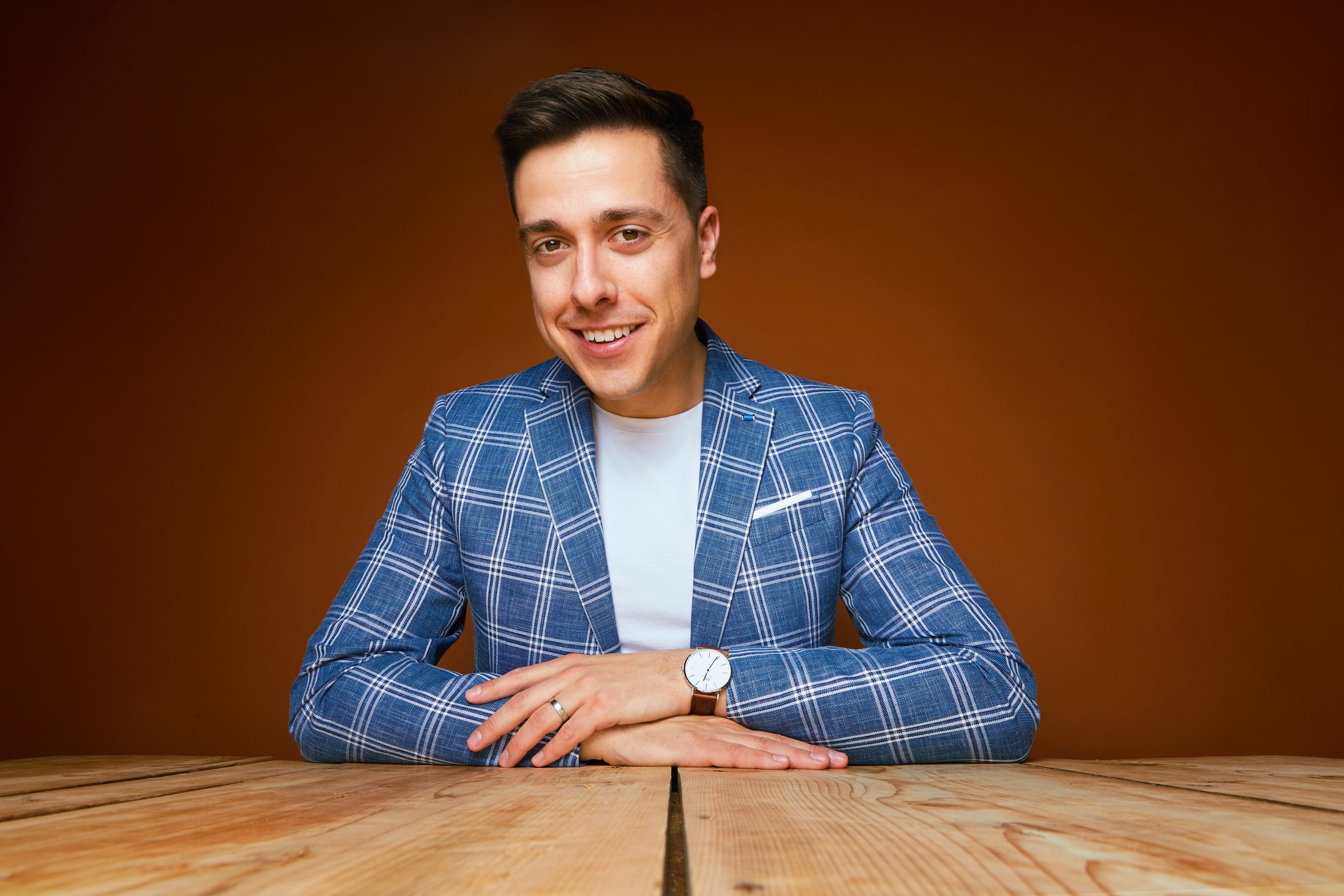 The same jacket and table against orange, hands folded, smiling