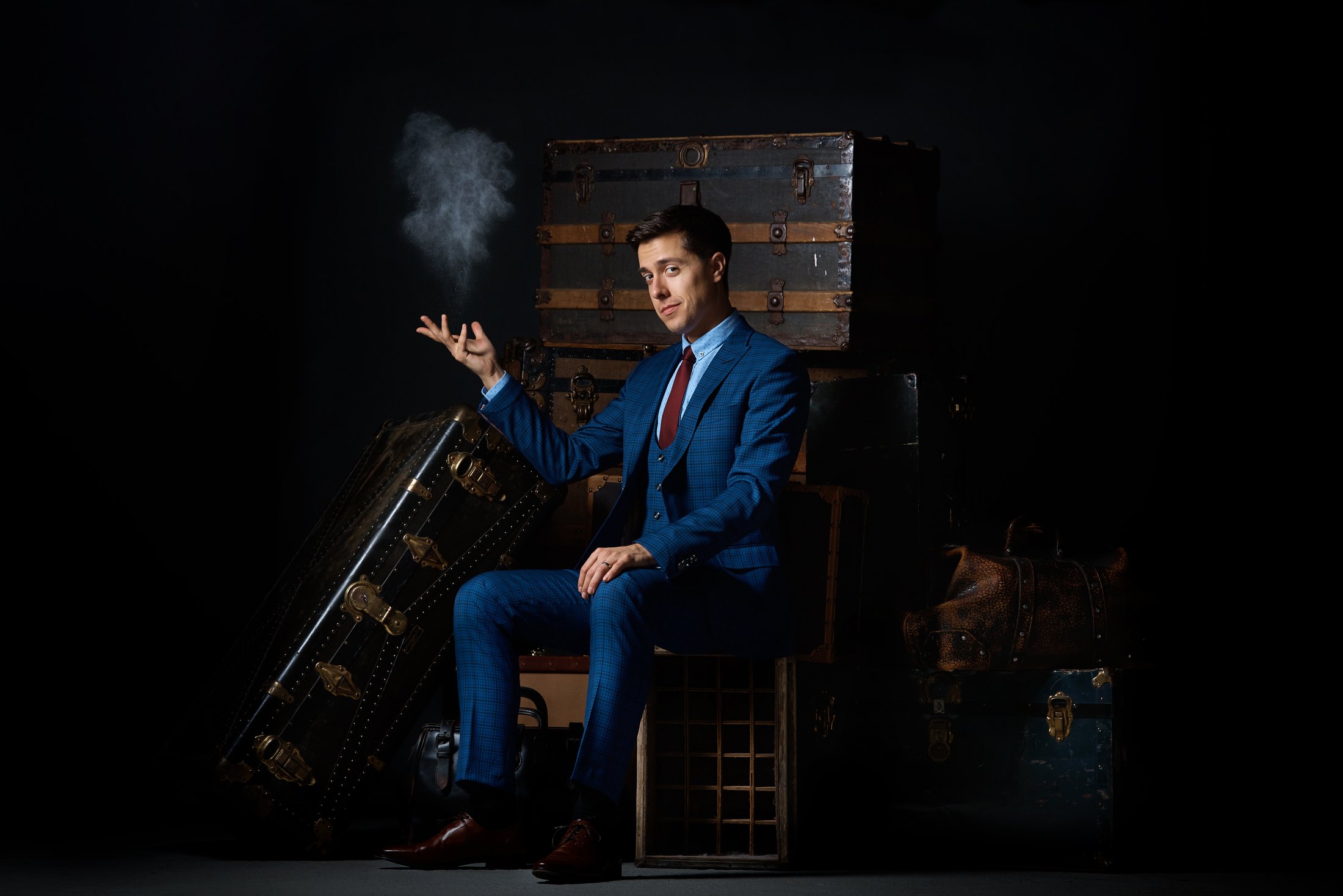 In a blue check three-piece suit among old steamer trunks, a puff of powder rising from his fingers