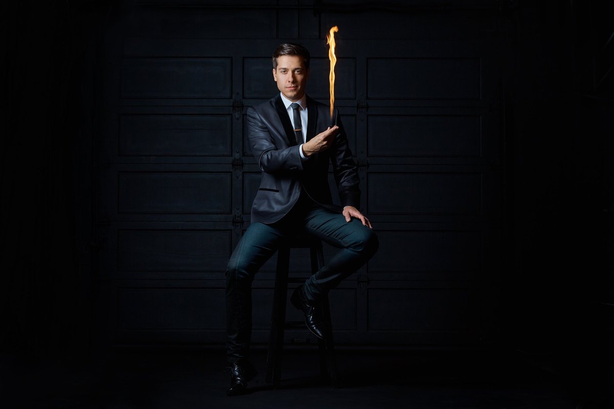 Sheldon Casavant on a stool against black panelled doors, a tall flame rising from his hand