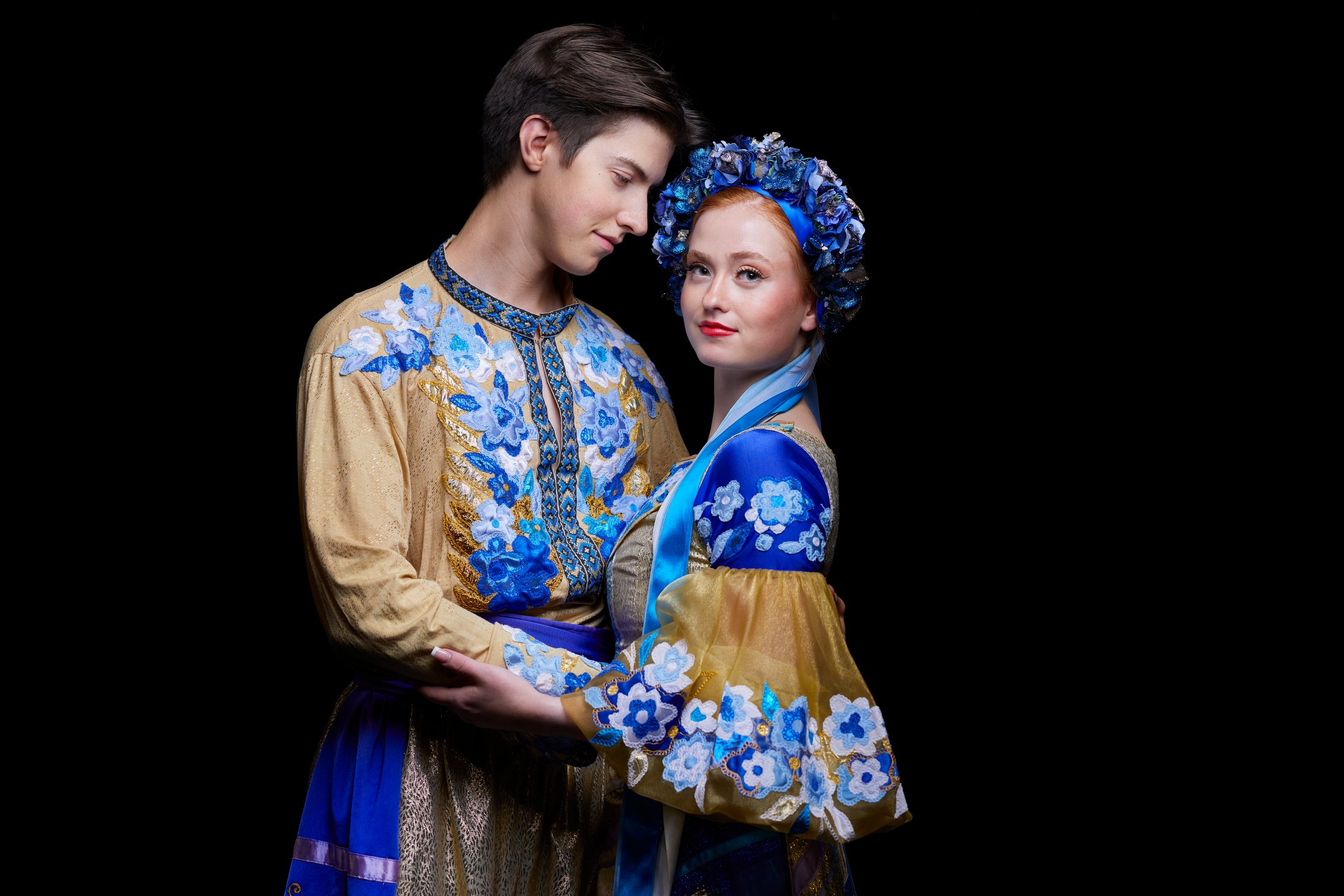 Two Shumka dancers in blue and white embroidered costumes, close together, against black