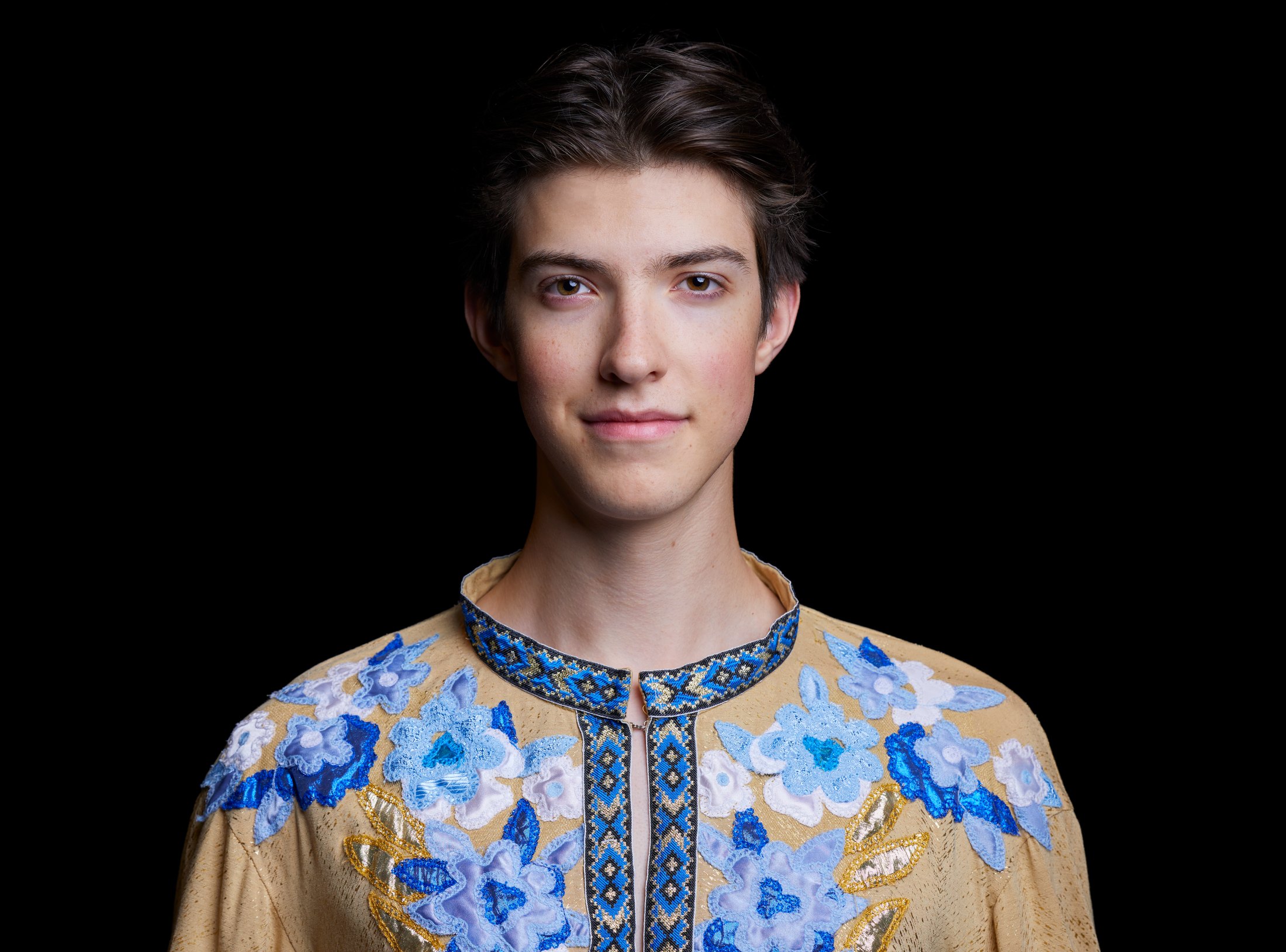 A young Shumka dancer in an embroidered blue and gold shirt, against black