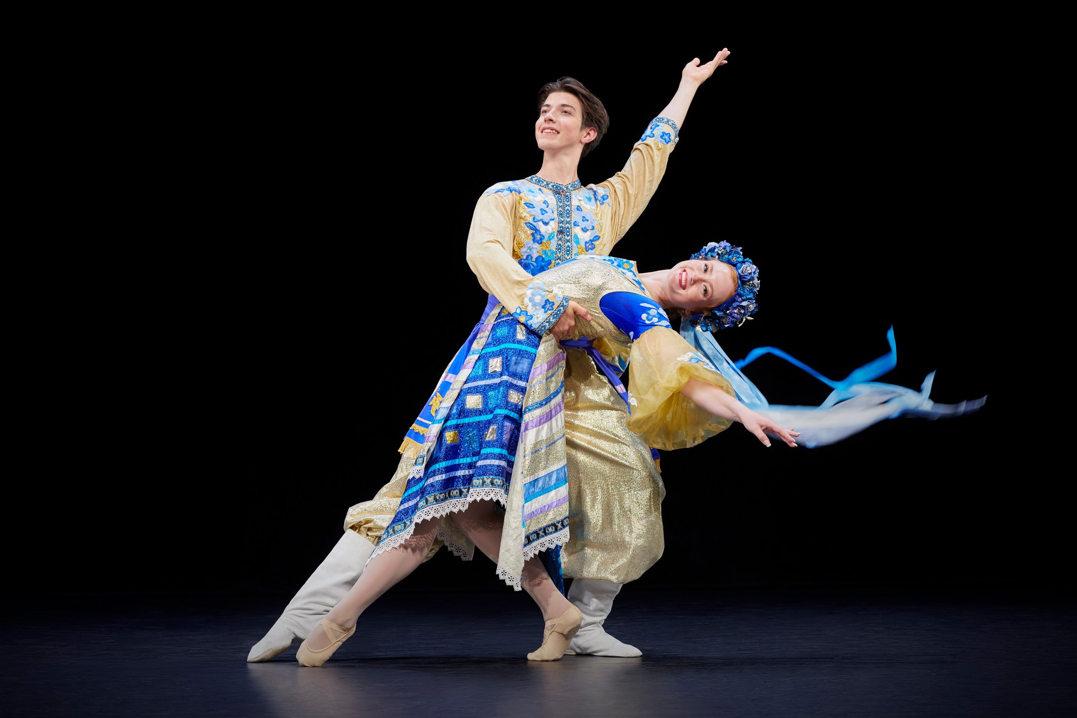 Two Shumka dancers in a lift, in blue and gold costumes, against black