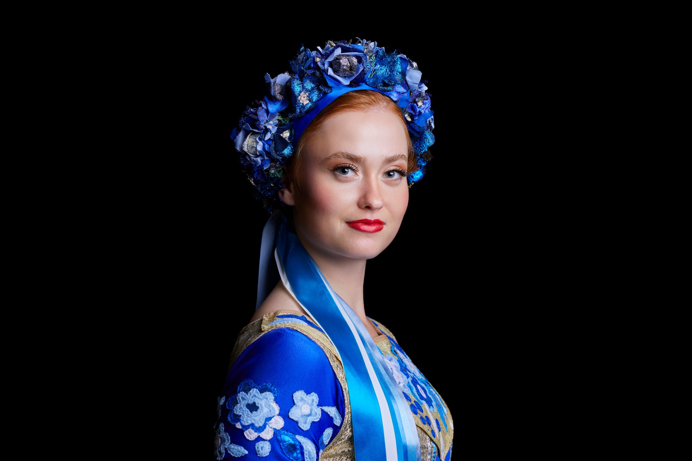 A Shumka dancer in a blue floral wreath and blue embroidered costume, against black