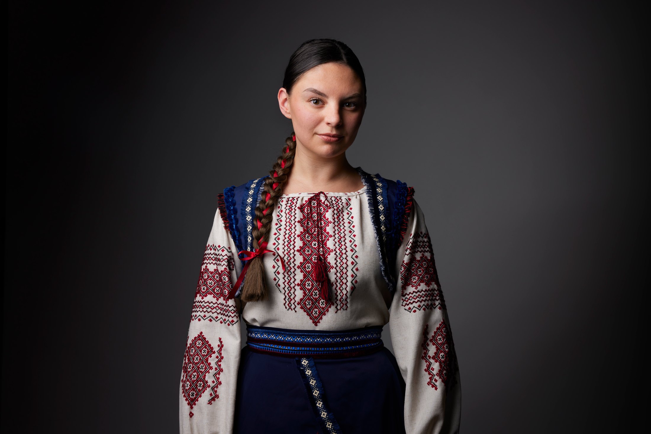 A dancer with a long braid in an embroidered blouse and dark vest, against grey