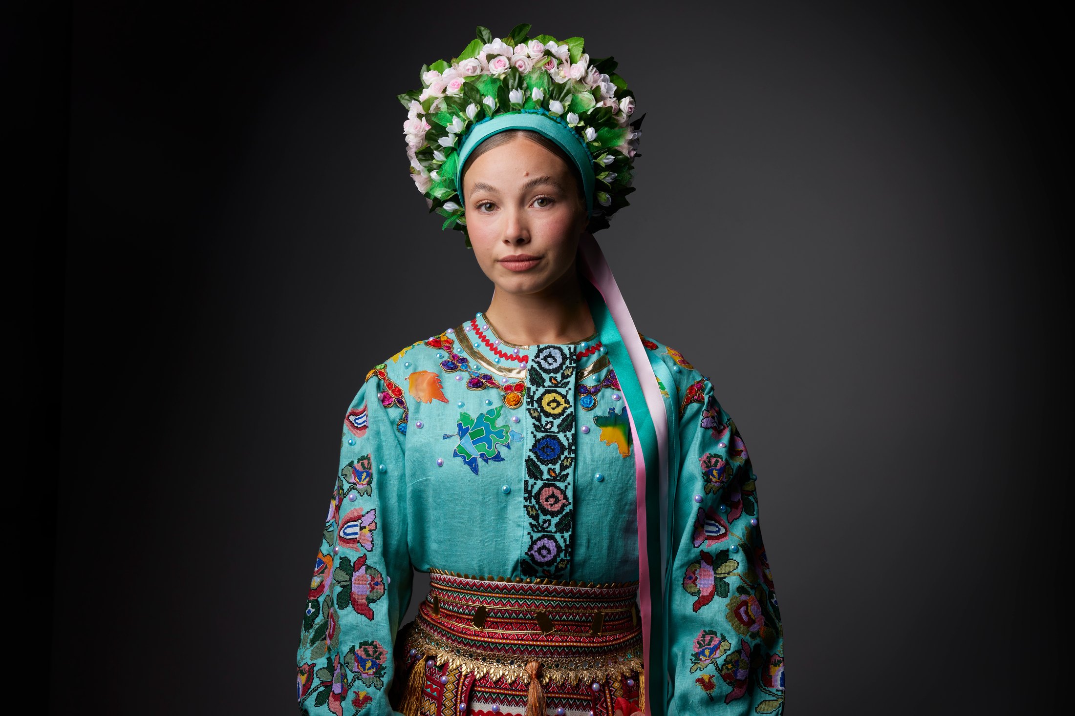 A dancer in a tall floral wreath and embroidered costume, against grey