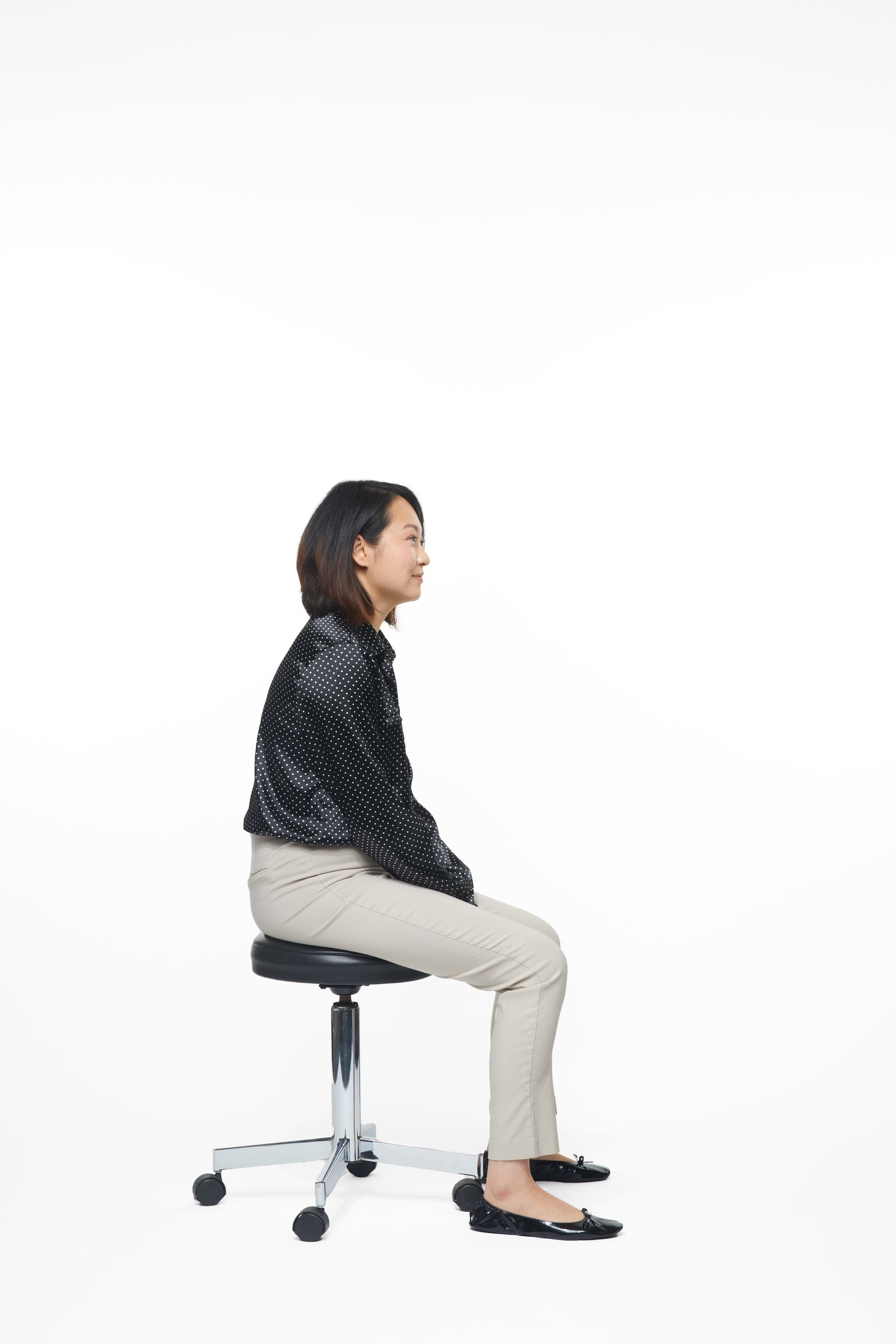 A woman in a black jacket and pale trousers seated on a stool, full length on white