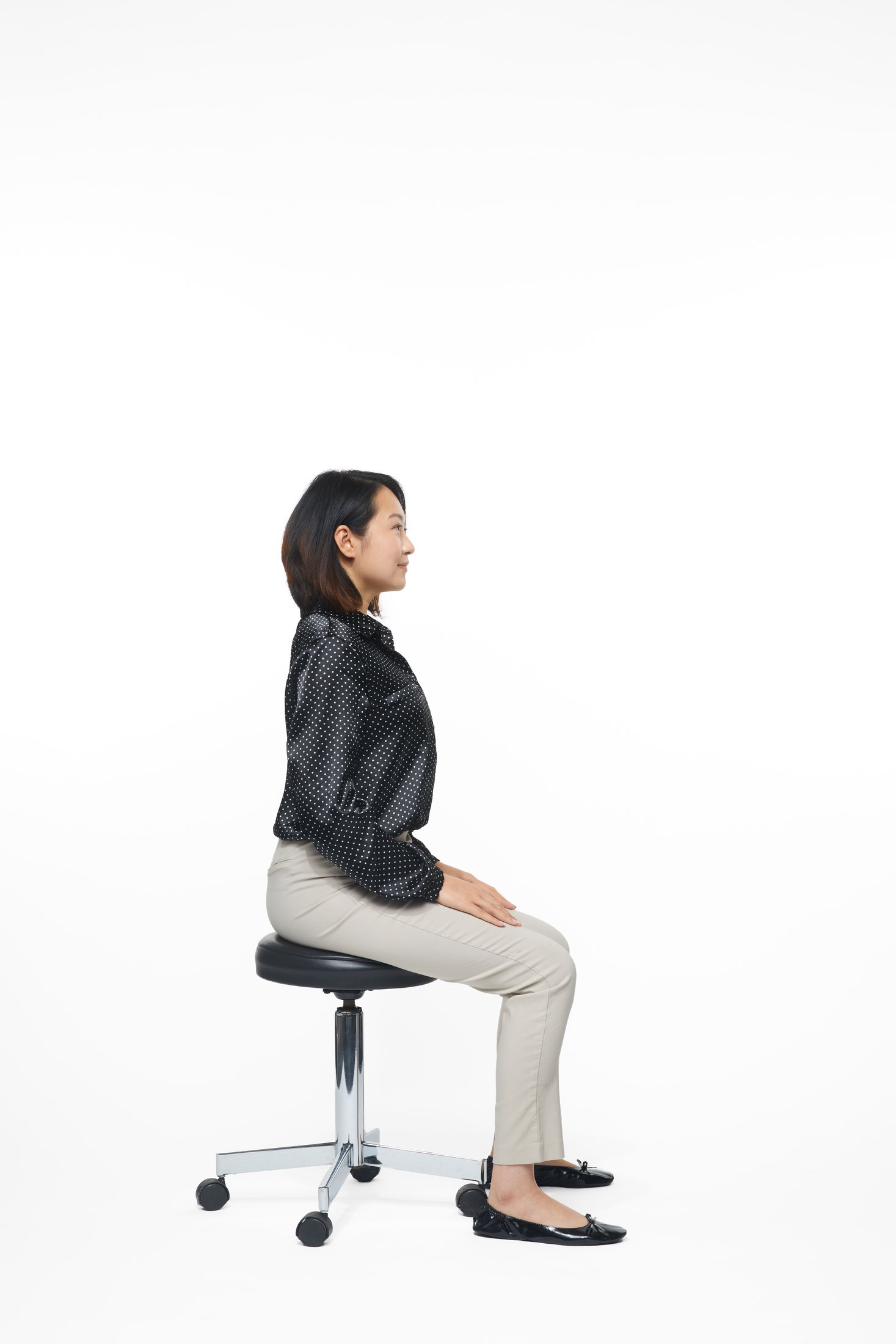 A woman in a black jacket and pale trousers seated on a stool, full length on white