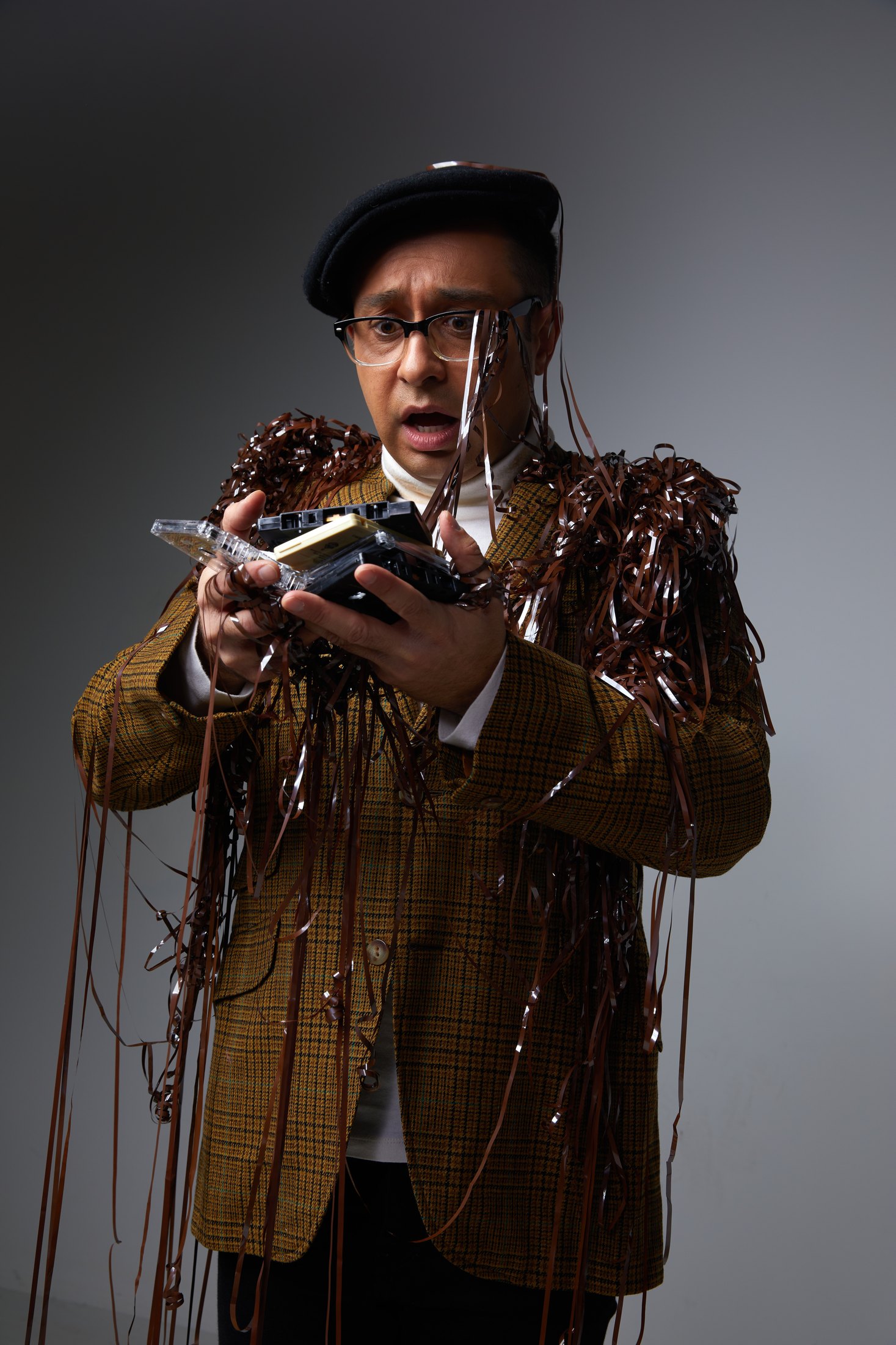A performer in a beret and tweed jacket, tangled in streamers of tape, against grey
