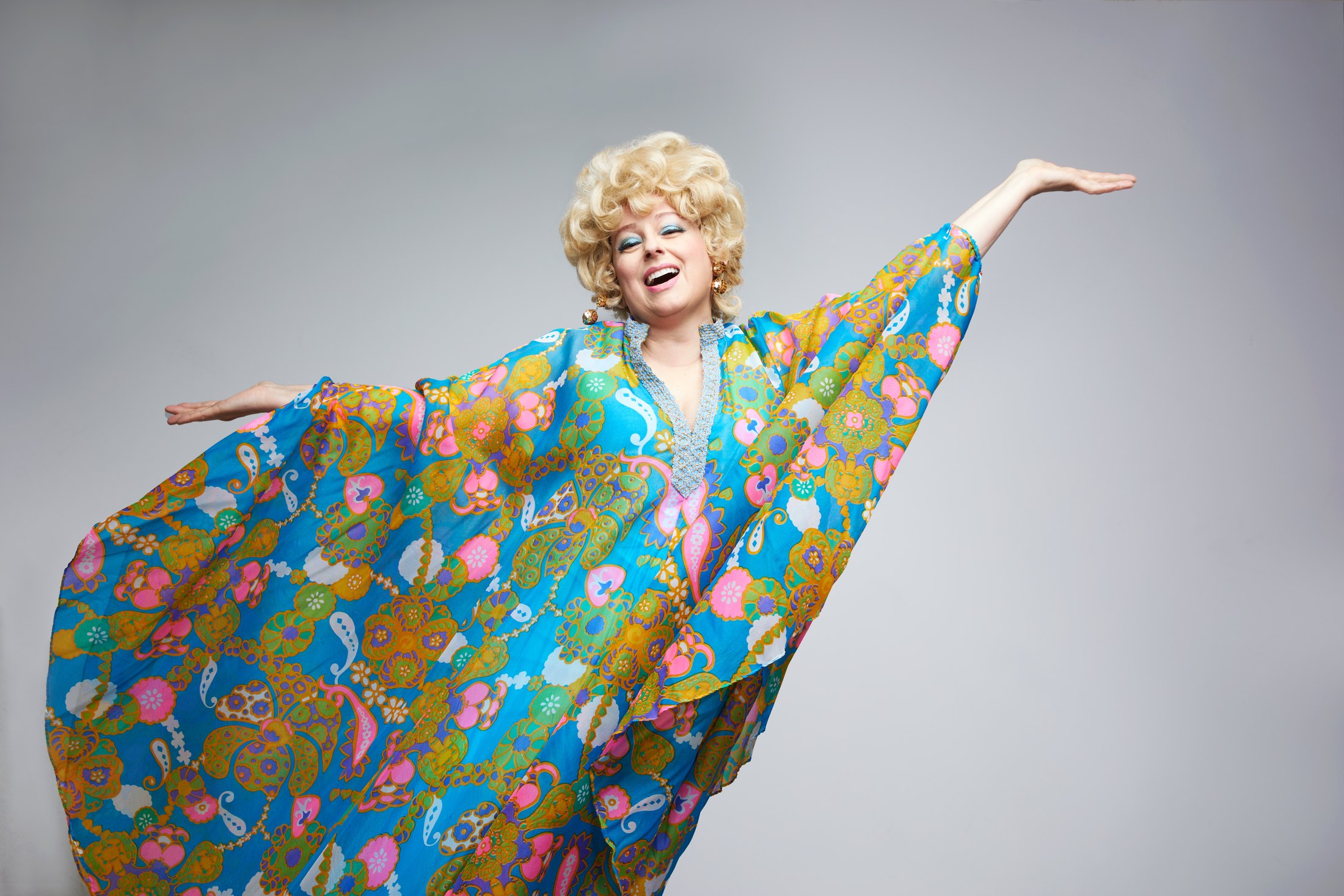 A performer in a blonde bouffant spreading the sleeves of a floral kaftan, against grey