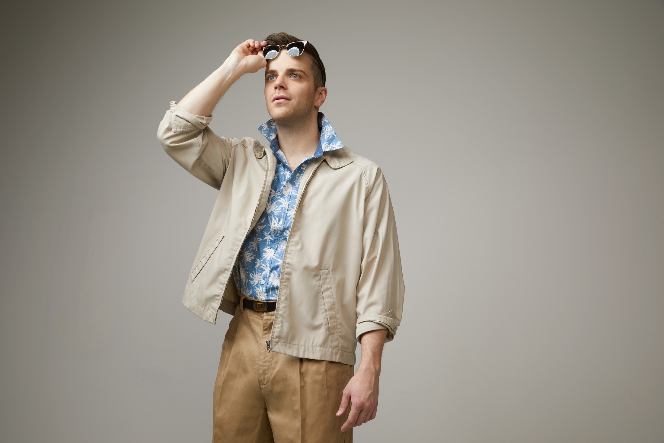 A performer in a beige jacket and patterned shirt, a hand at his sunglasses, against grey