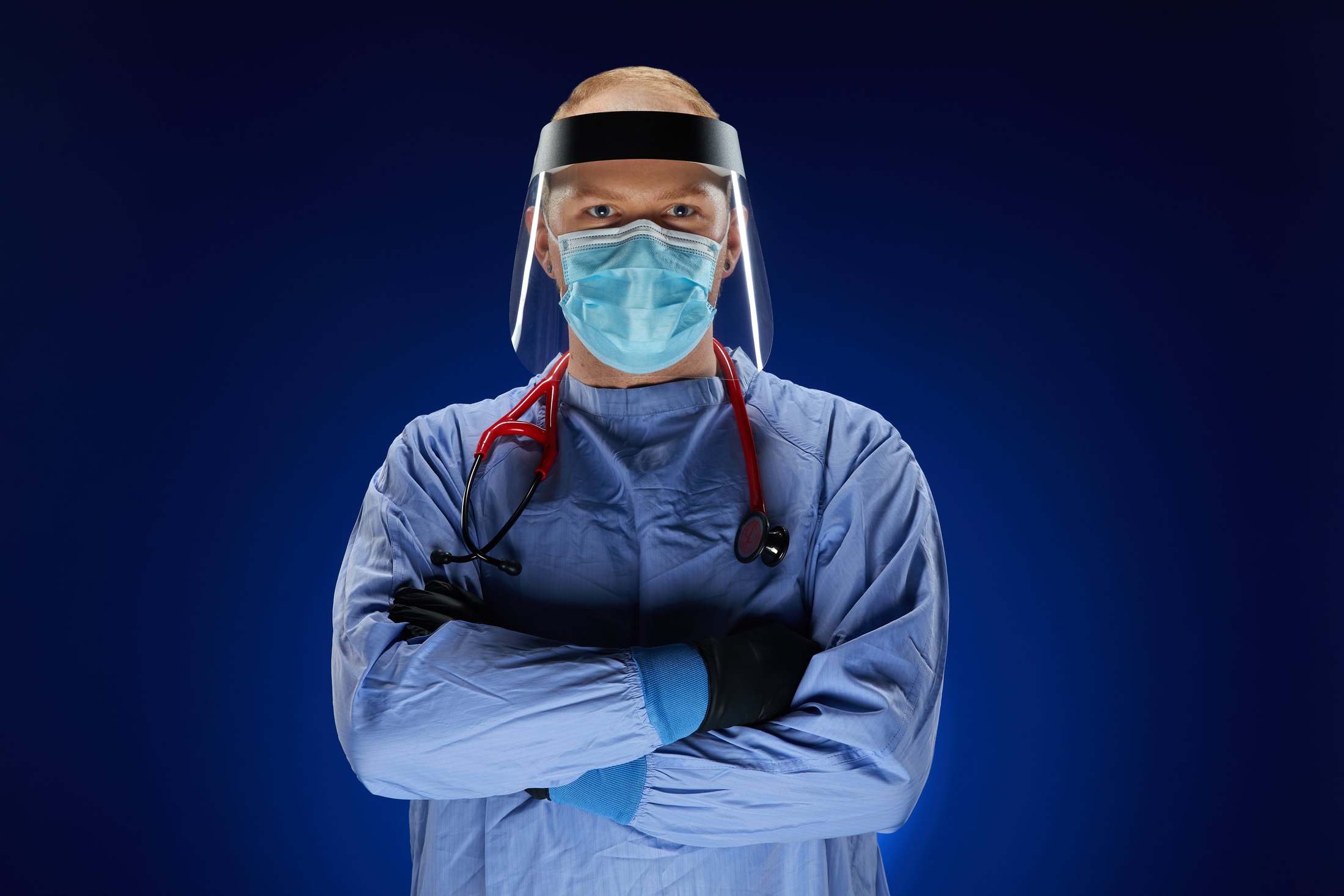 A nurse in a blue isolation gown, face shield and mask, arms crossed, against deep blue