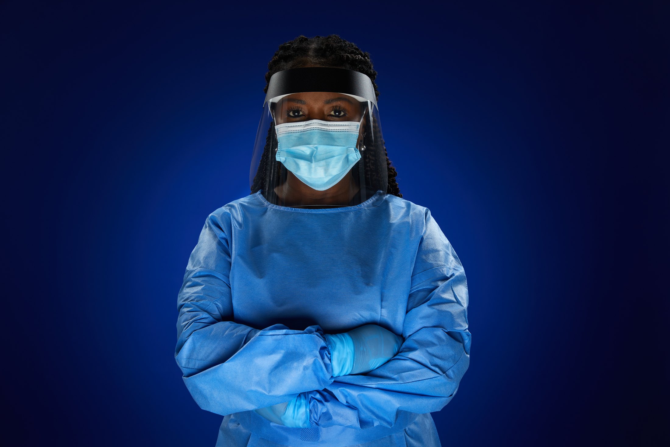 A nurse in a blue isolation gown, face shield and mask, arms crossed, against deep blue