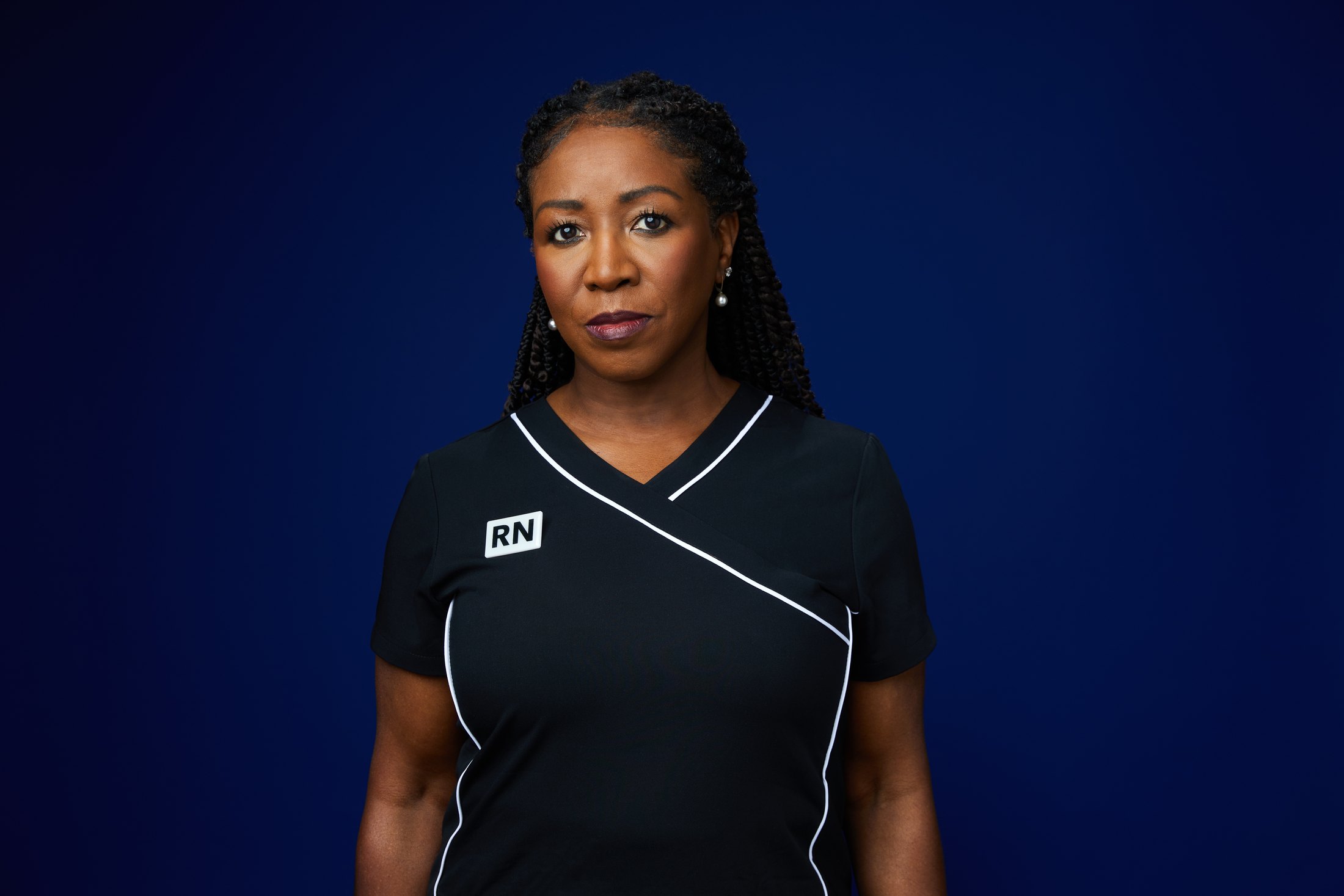A nurse in black scrubs with white piping, against deep blue