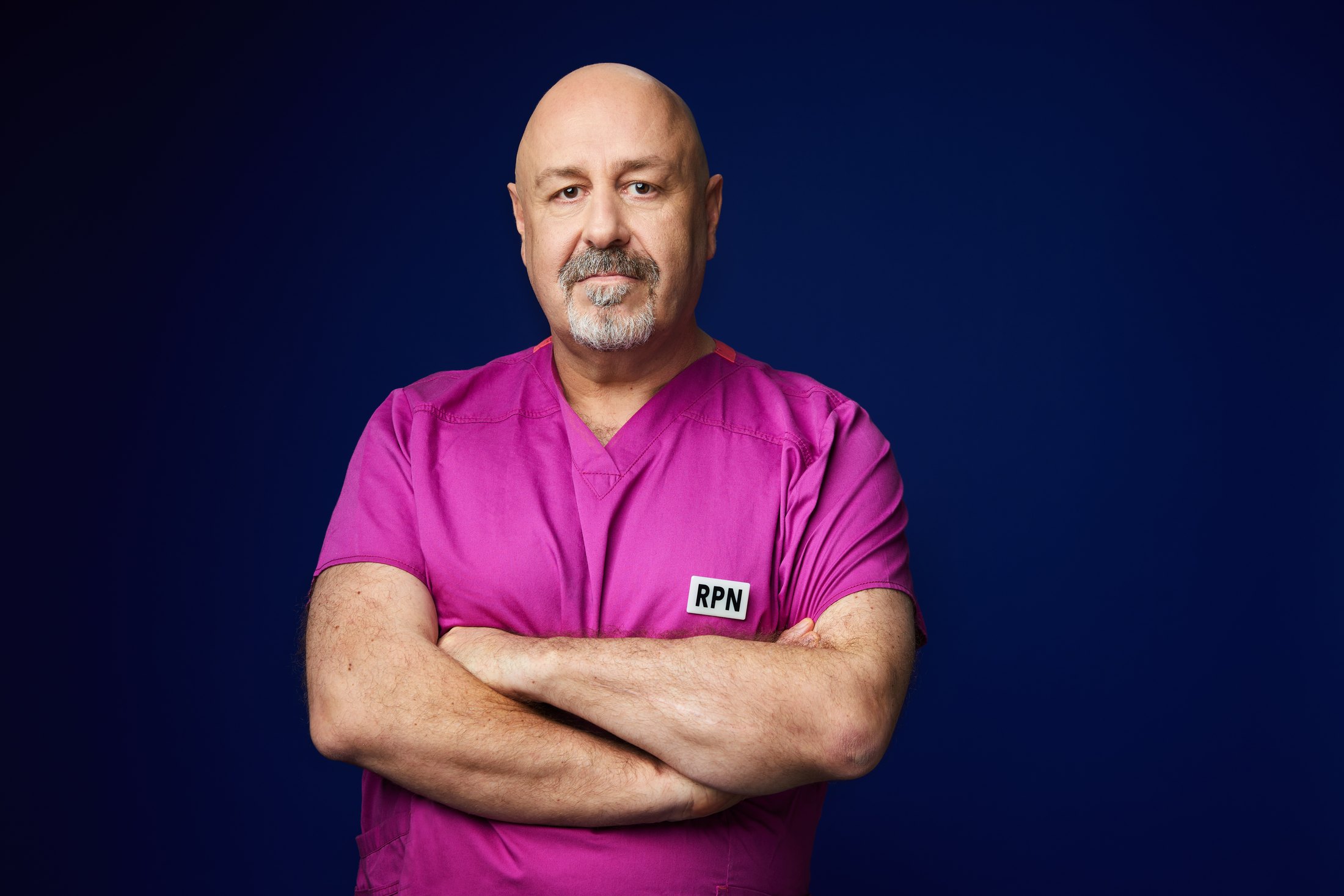 A nurse in magenta scrubs, arms crossed, against deep blue