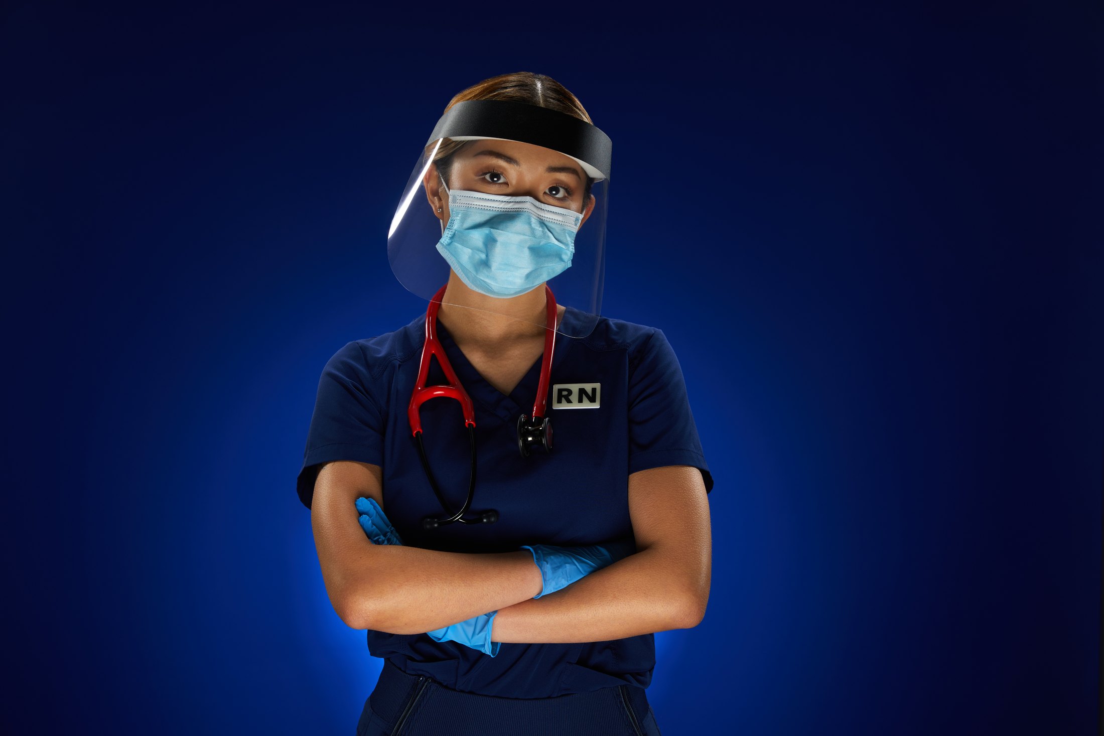 A nurse in navy scrubs and blue gloves, face shield and mask, a red stethoscope at her neck