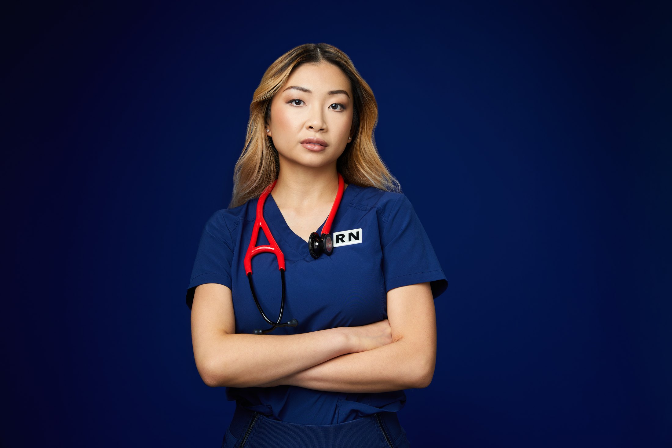 A nurse in navy scrubs with a red stethoscope, arms crossed, against deep blue