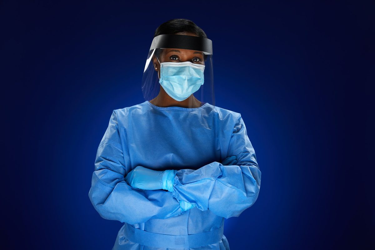 A nurse rim-lit in a blue isolation gown, face shield and mask, arms crossed
