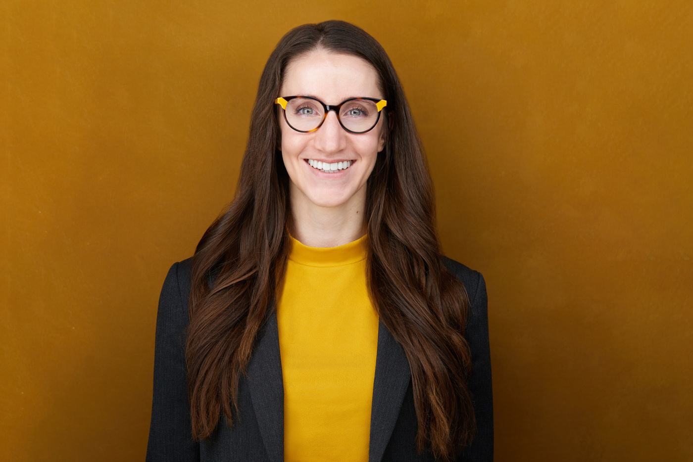 Her business headshot: glasses, a mustard top and a charcoal blazer against gold