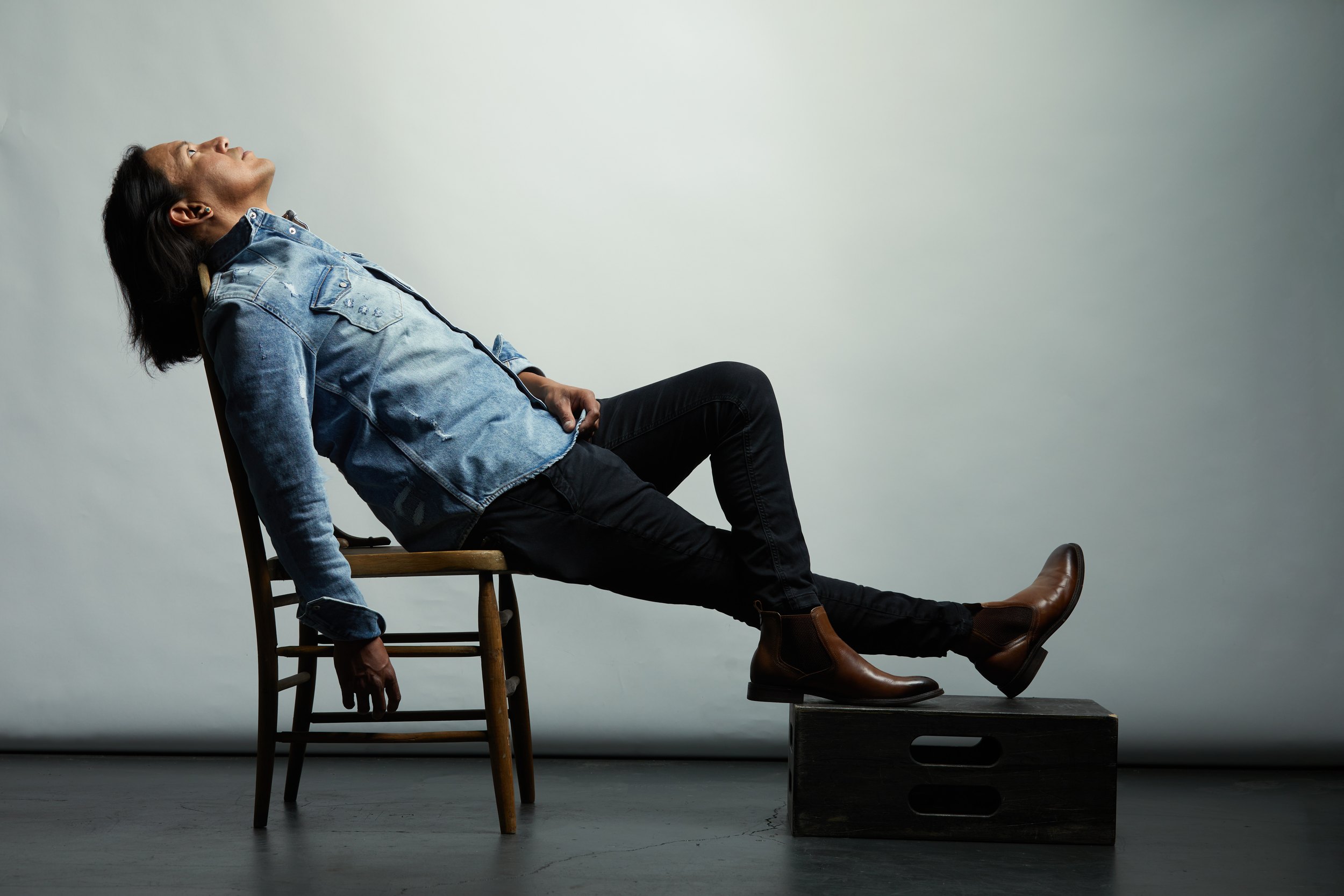 The same session, leaning right back in a wooden chair with the boots up on a crate, on white