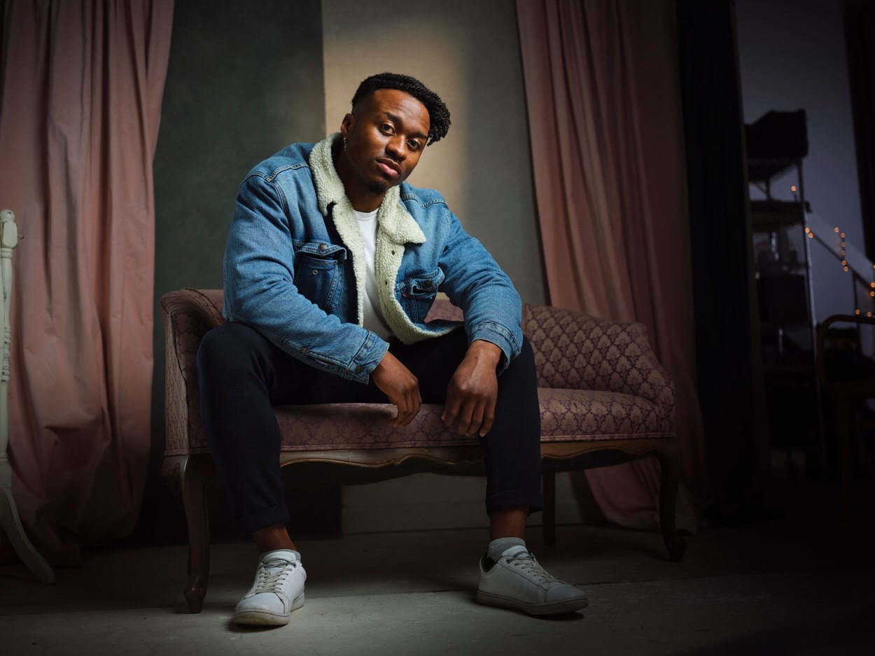 The same session, a denim jacket, seated on a velvet settee between curtains