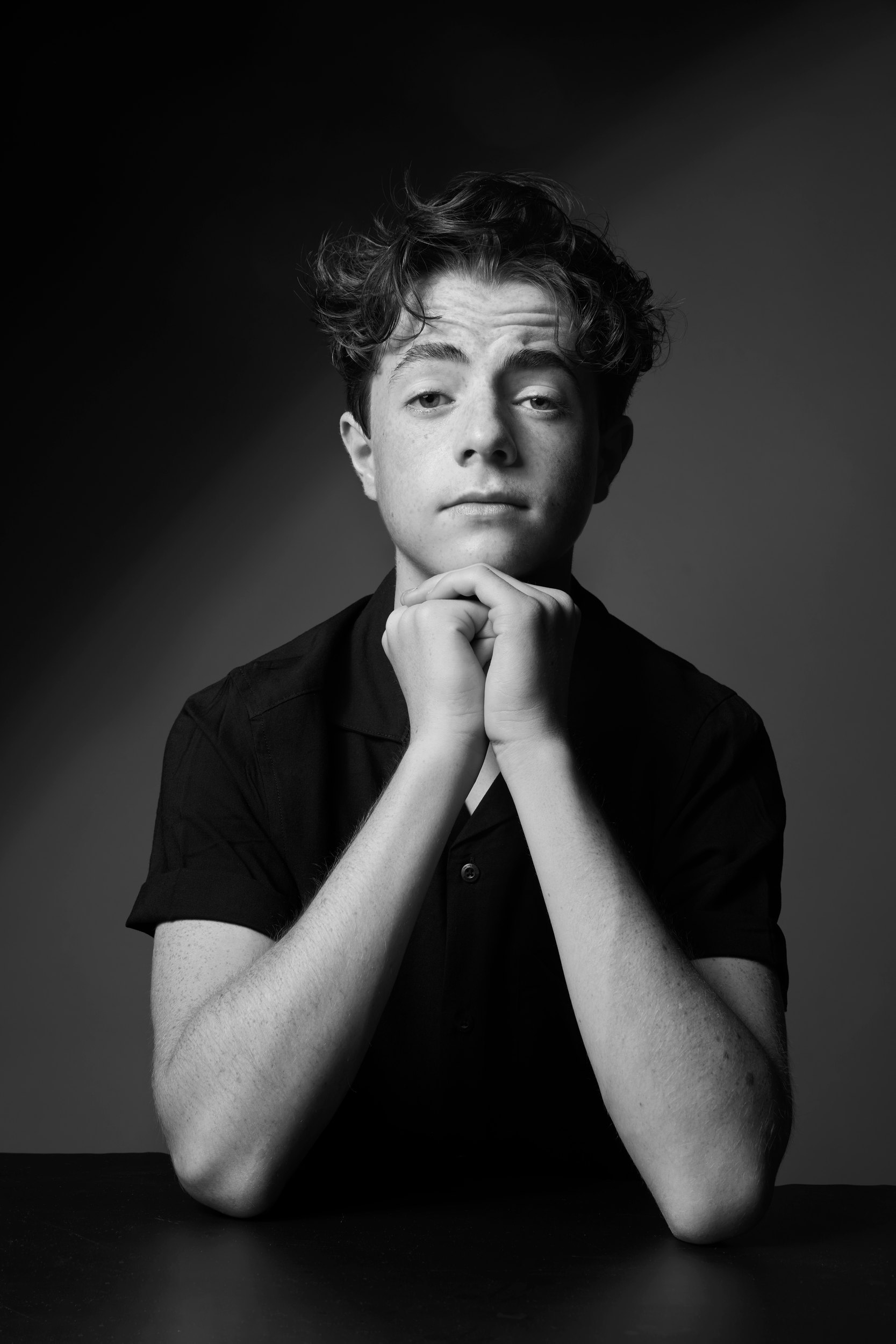 In black and white, a black polo, chin on his hands